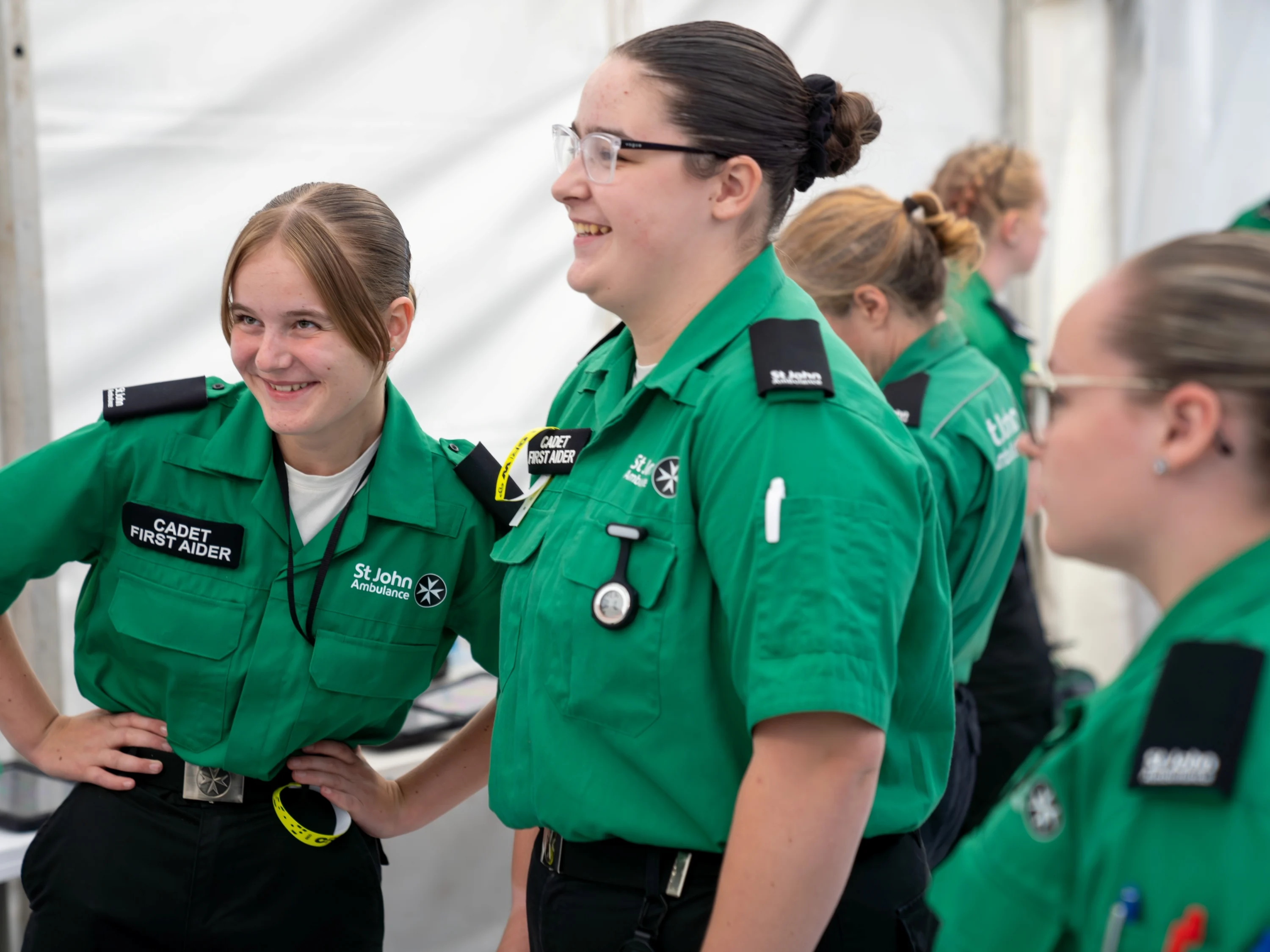 A group of Cadet First Aiders standing around in a white tent. Two are smiling with their hands on their hips in front of another volunteer.