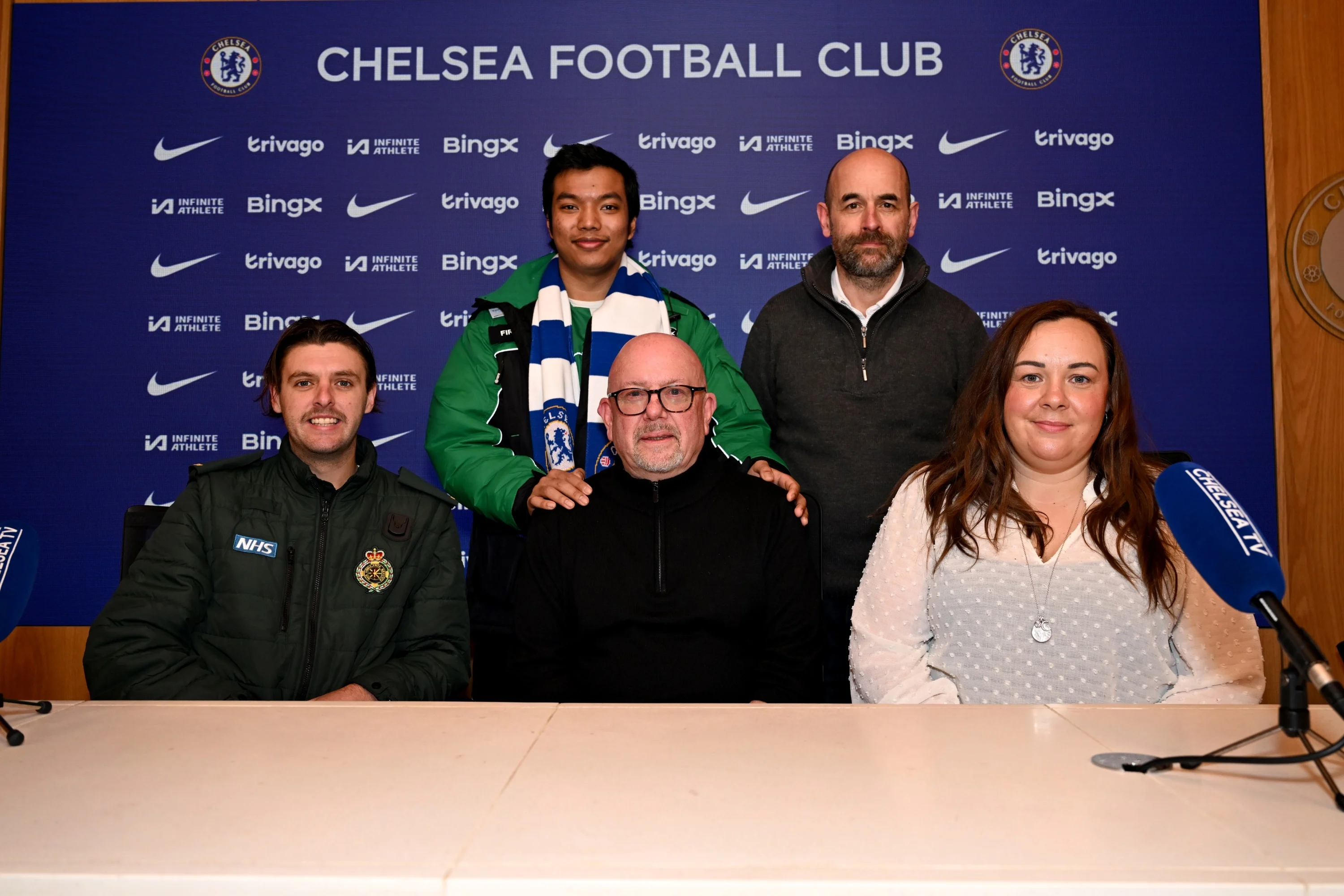 Paul Archer sat at a table with the Chelsea Football Club logo behind him surrounded by the medical team who saved his life. 