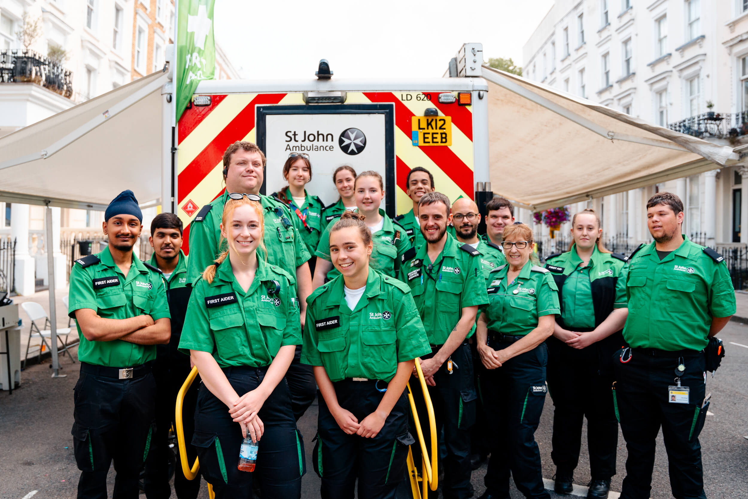 A photograph of a group of St John volunteers smiling. They are outdoors and standing in front of a St John ambulance.