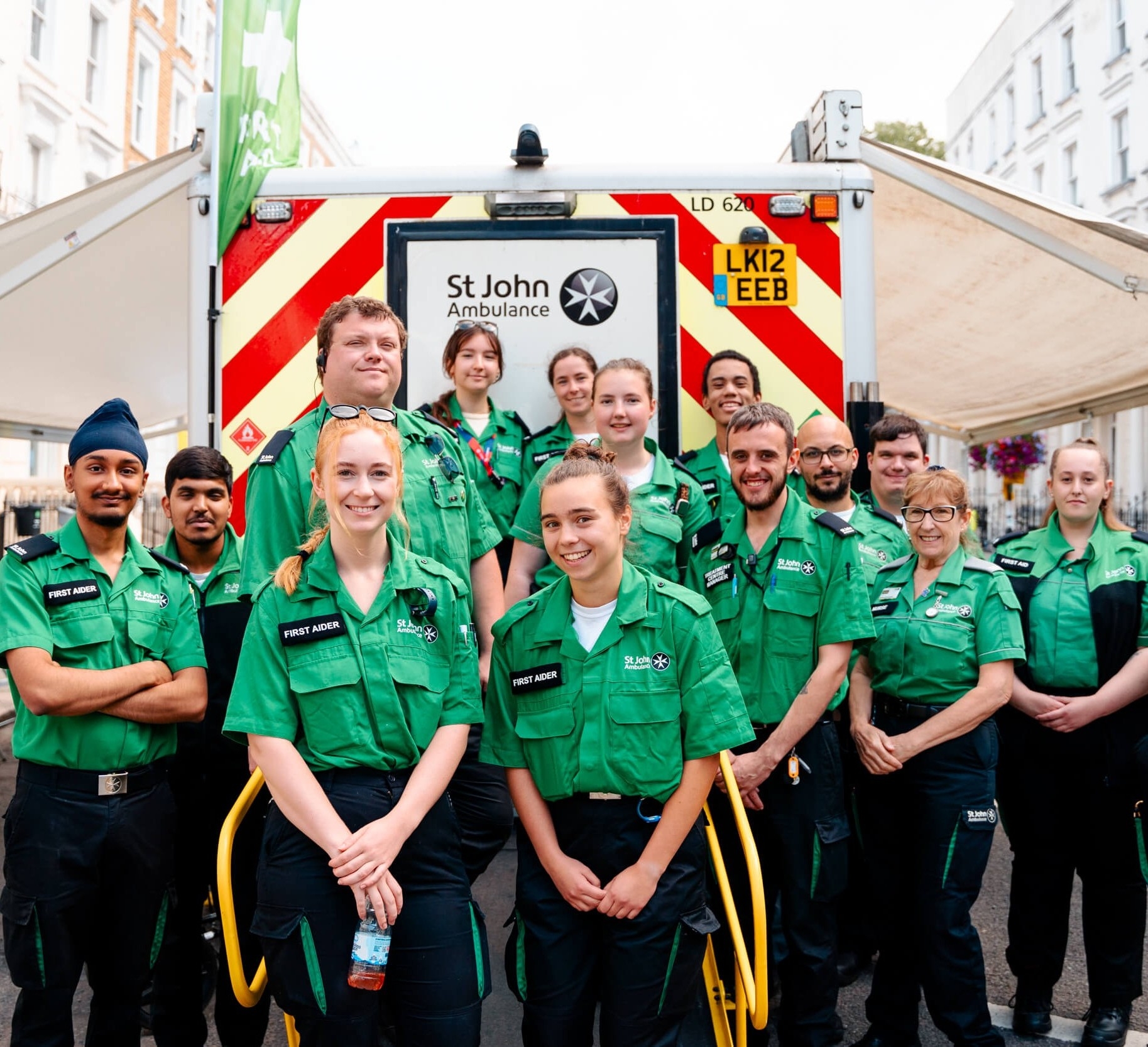 A photograph of a group of St John volunteers smiling. They are outdoors and standing in front of a St John ambulance.