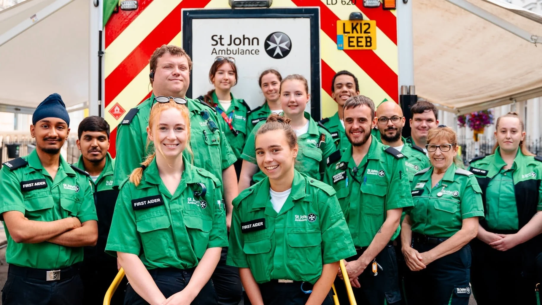 A photograph of a group of St John volunteers smiling. They are outdoors and standing in front of a St John ambulance.