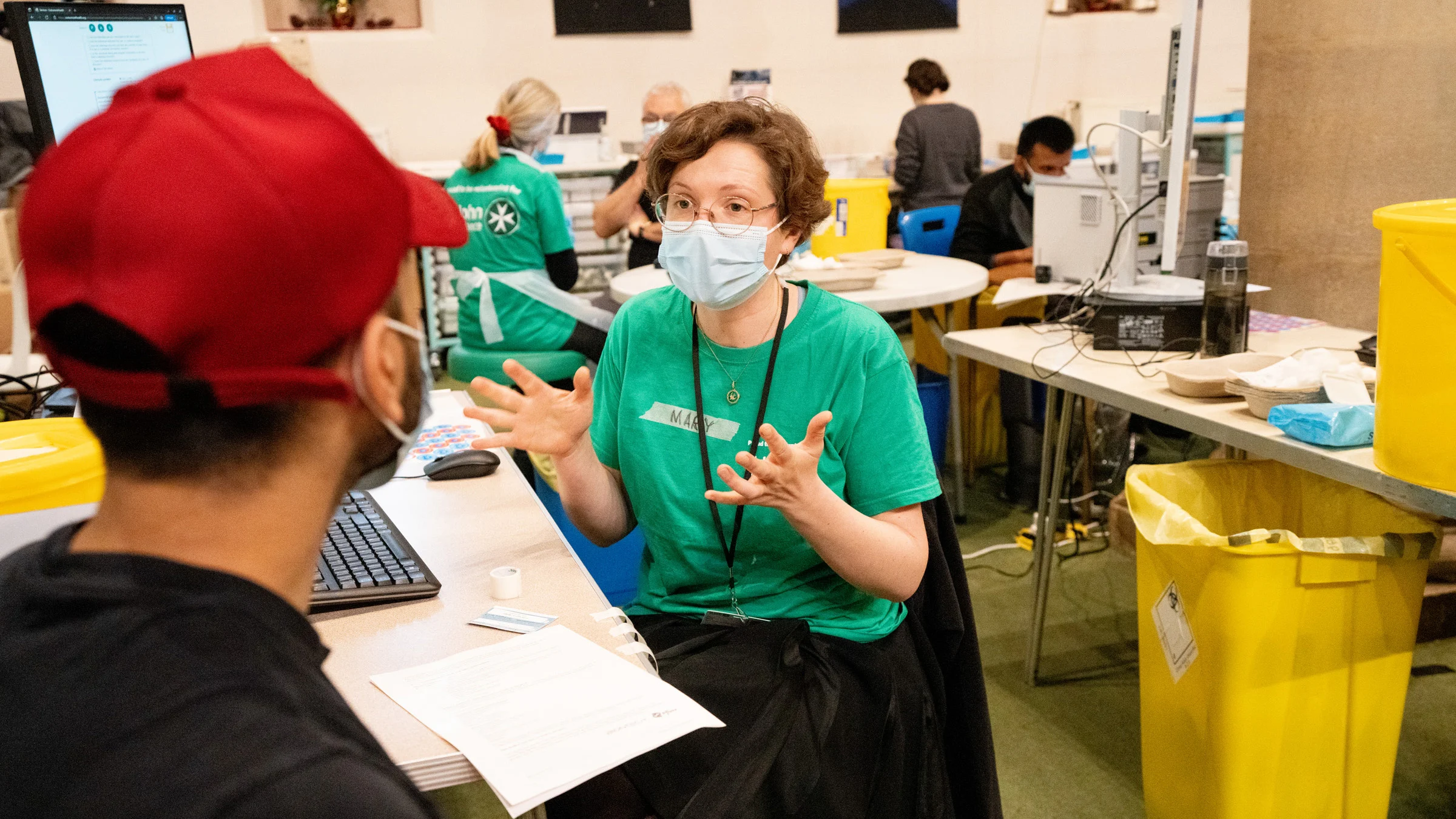 A photograph of Mary talking to a patient in a vaccination centre. She is wearing a St John Ambulance t-shirt and a face mask.