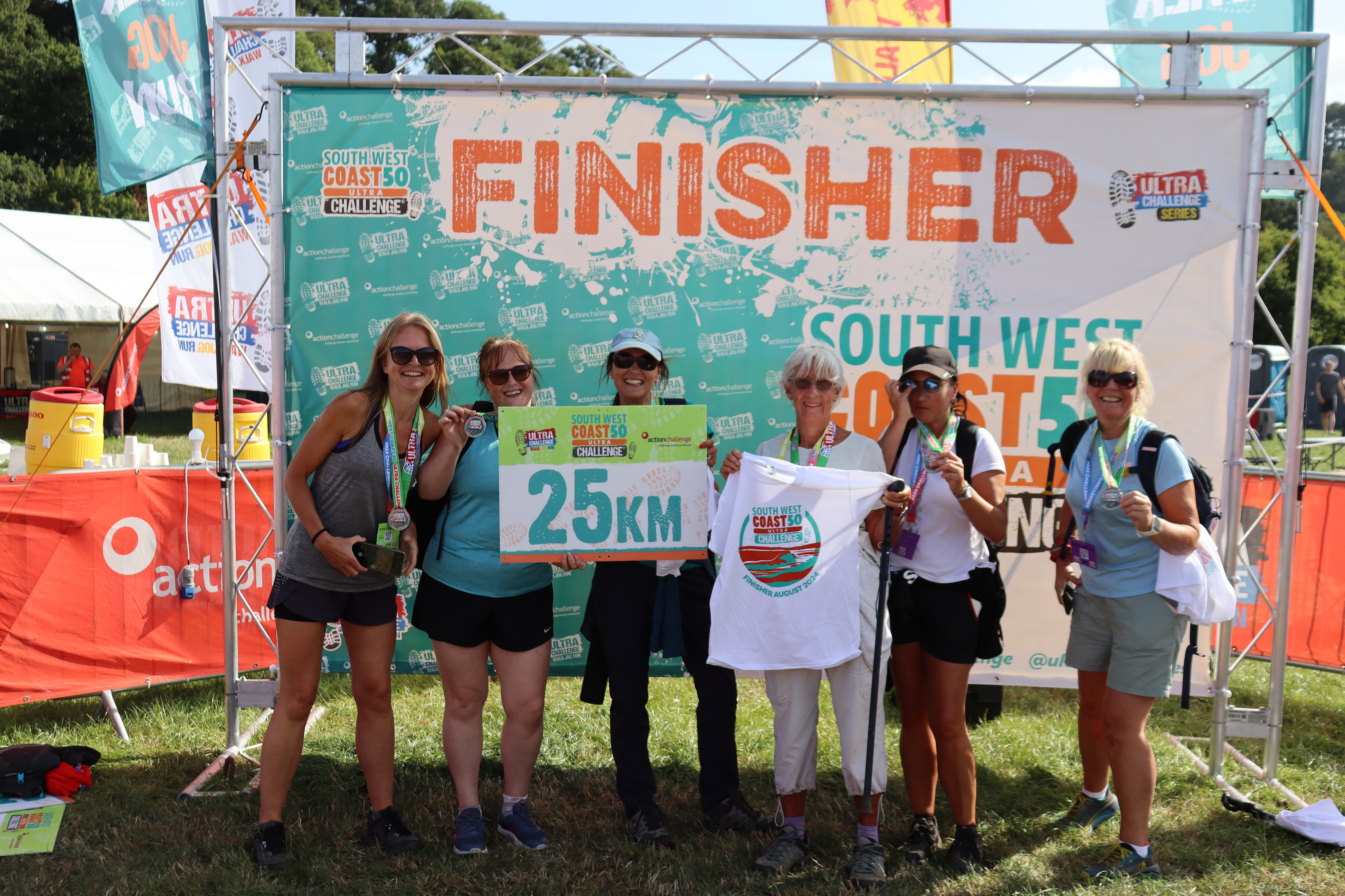 Six people stood in front of a large board saying 'Finisher' at the South West 50 Ultra Challenge finish line. They are holding up a t-shirt and a sign that says '25km'. 