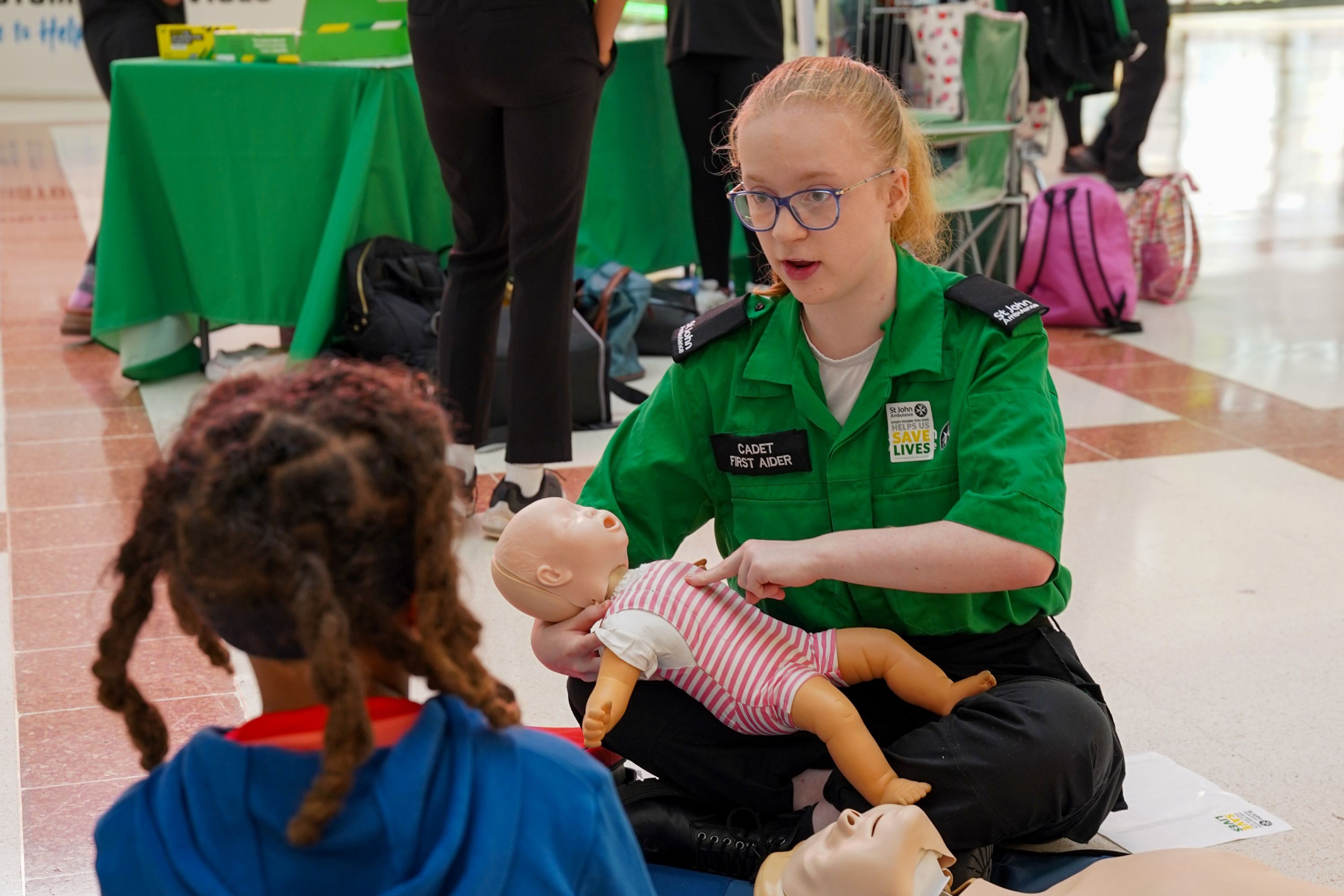 A St John Volunteer demonstrating baby first aid to a toddler, using a baby manikin.