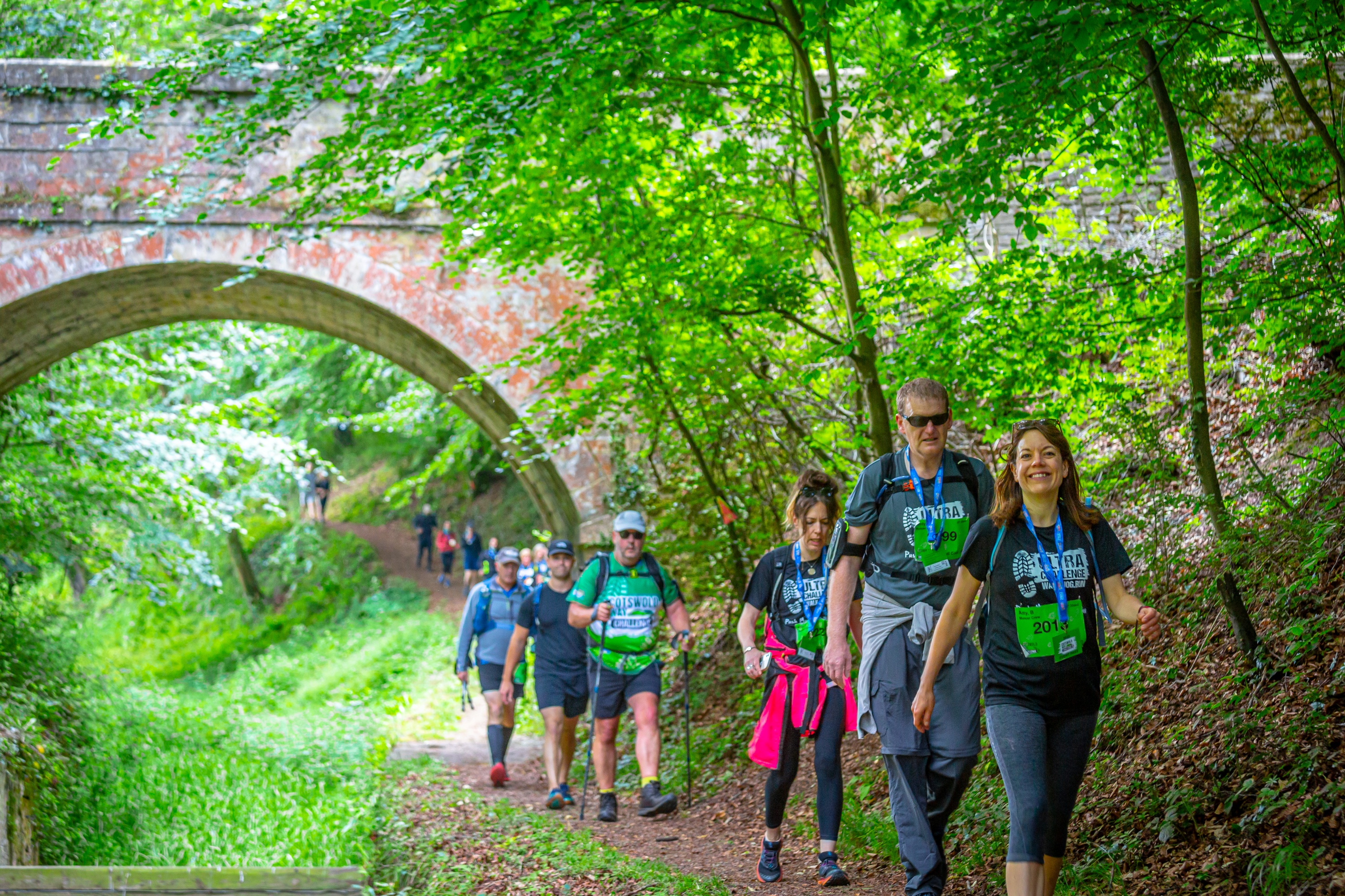 A line of people walking under a bridge and along a woodland footpath. They are wearing t-shirts with the Cotswold Way Ultra Challenge logo on it.