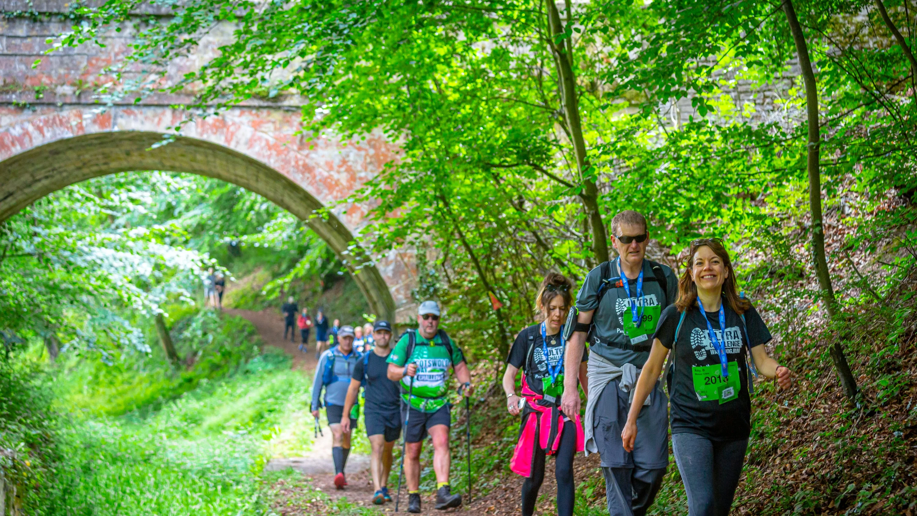 A line of people walking under a bridge and along a woodland footpath. They are wearing t-shirts with the Cotswold Way Ultra Challenge logo on it.
