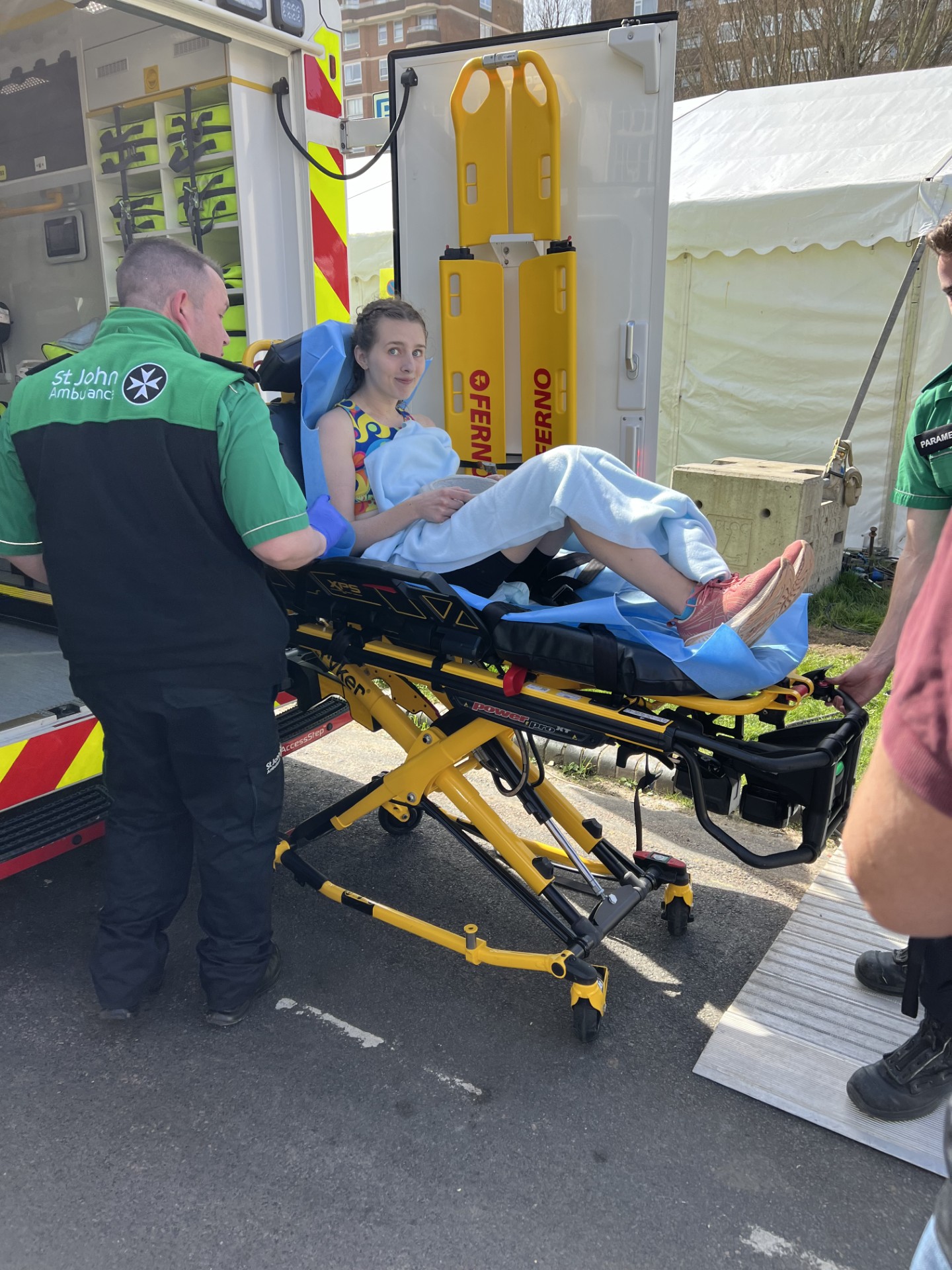 A photograph of Holly on a stretcher. She is wearing running gear and covered in a light sheet. A St John volunteer is attending to her.