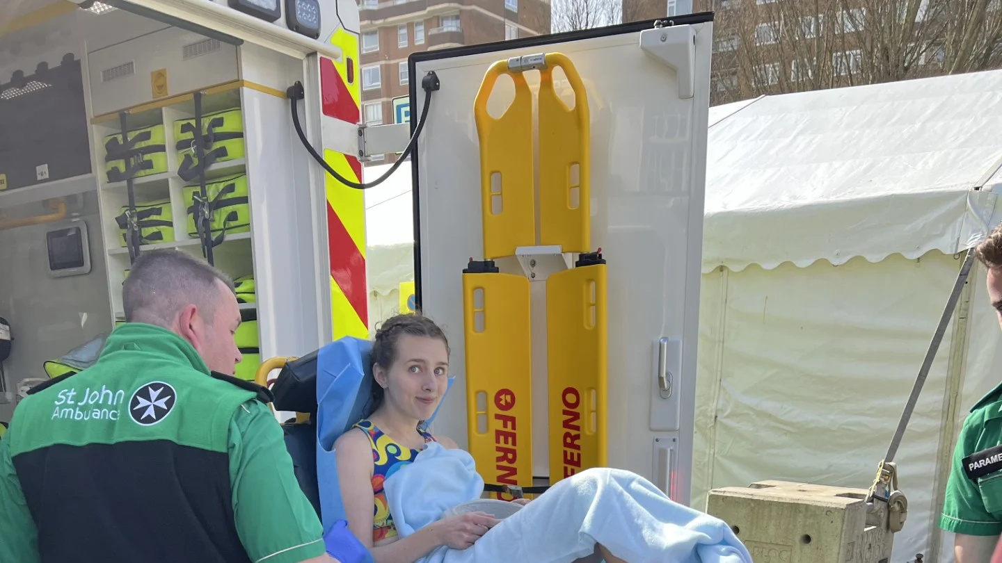 A photograph of Holly on a stretcher. She is wearing running gear and covered in a light sheet. A St John volunteer is attending to her.