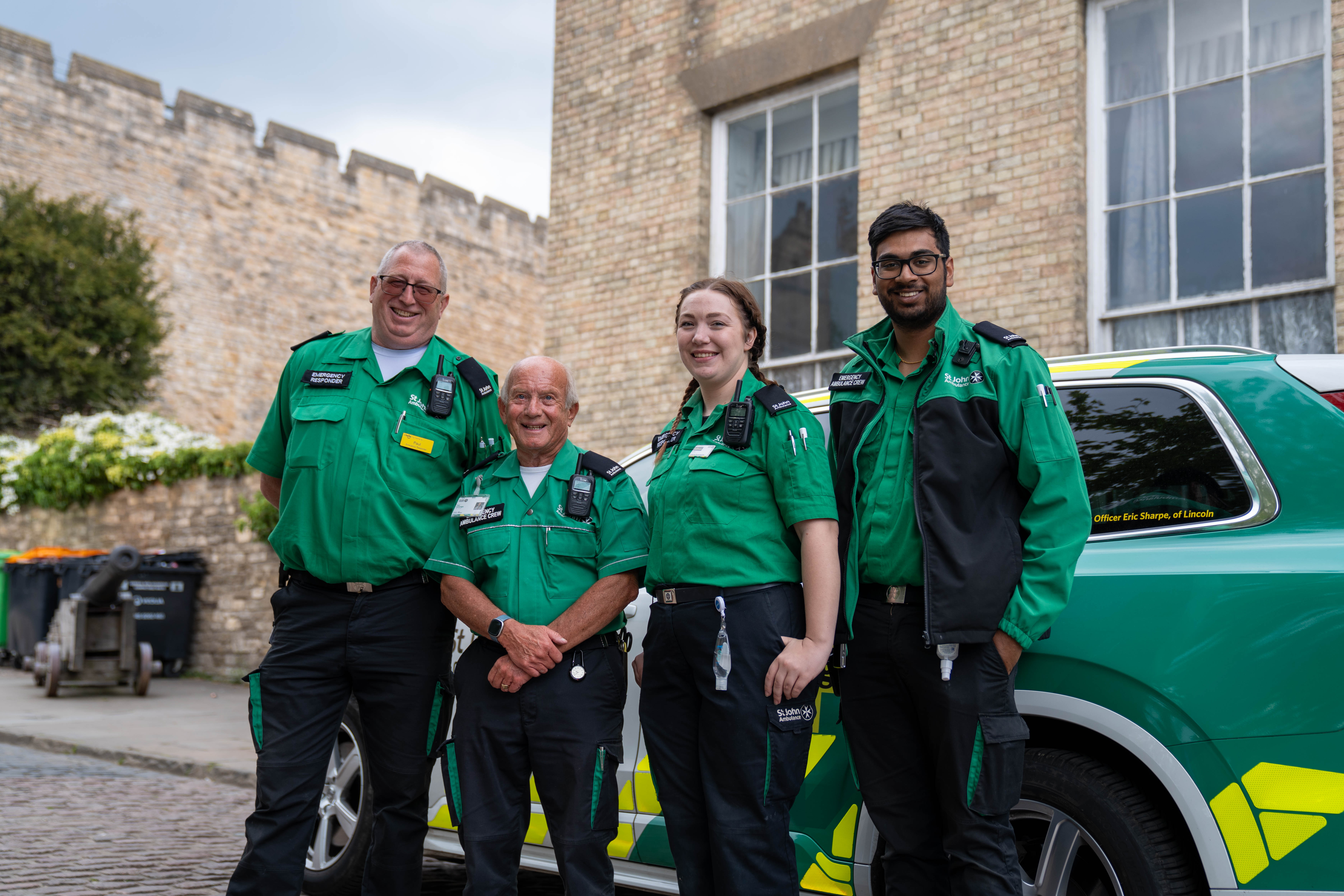 Four St John Ambulance volunteers stood smiling in front of an emergency responder vehicle. 