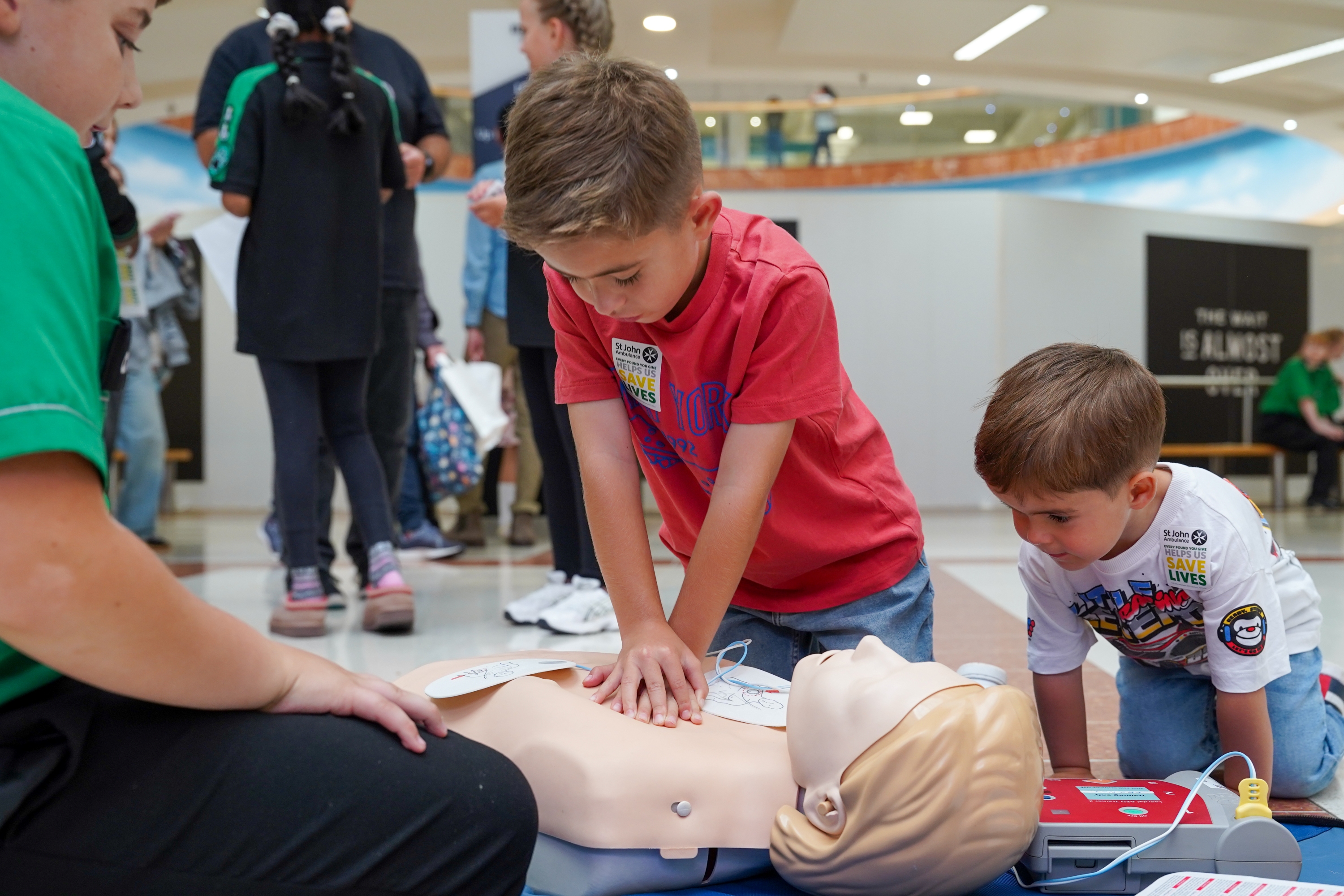 A photograph of a first aid demonstration in a shopping centre. A young boy performs chest compressions on a Manikin as a younger boy watches on.