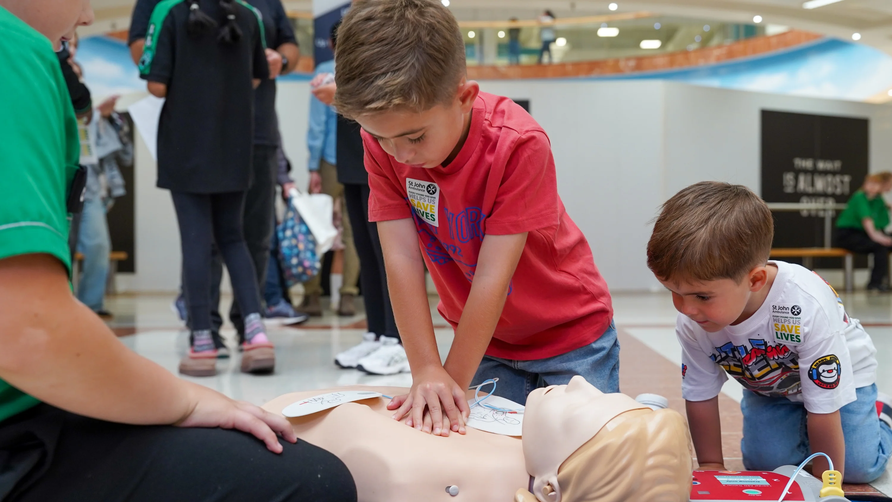 A photograph of a first aid demonstration in a shopping centre. A young boy performs chest compressions on a Manikin as a younger boy watches on.