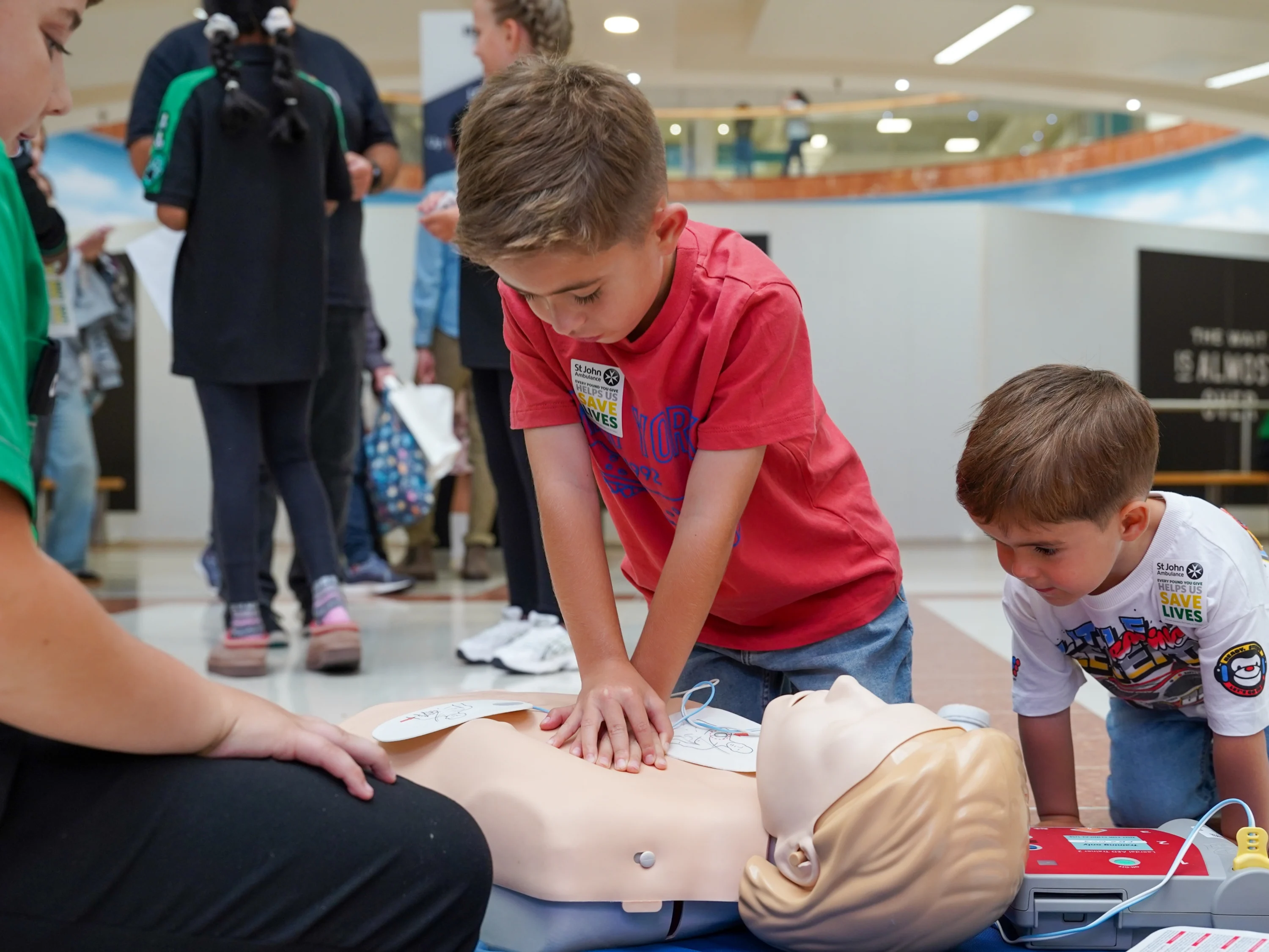 A photograph of a first aid demonstration in a shopping centre. A young boy performs chest compressions on a Manikin as a younger boy watches on.