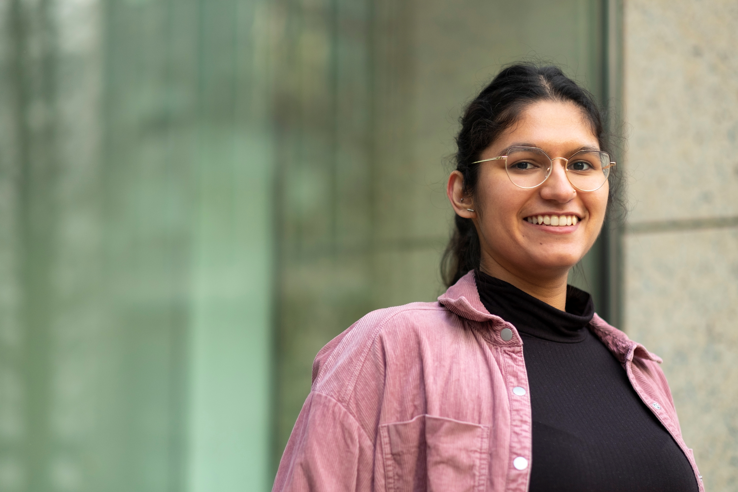 A photograph of Mausum smiling at the camera. She is wearing glasses and a pink overshirt.