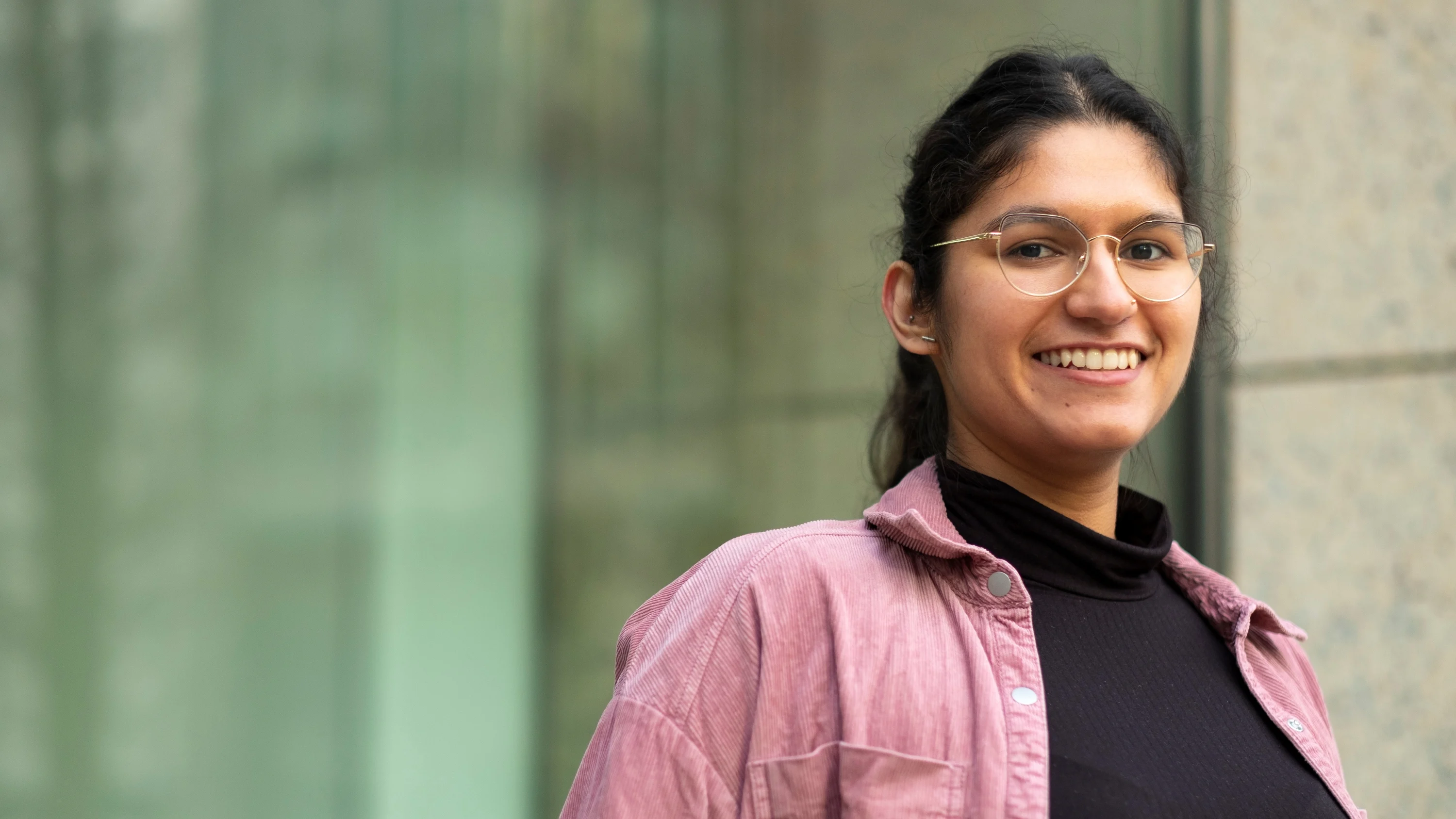 A photograph of Mausum smiling at the camera. She is wearing glasses and a pink overshirt.