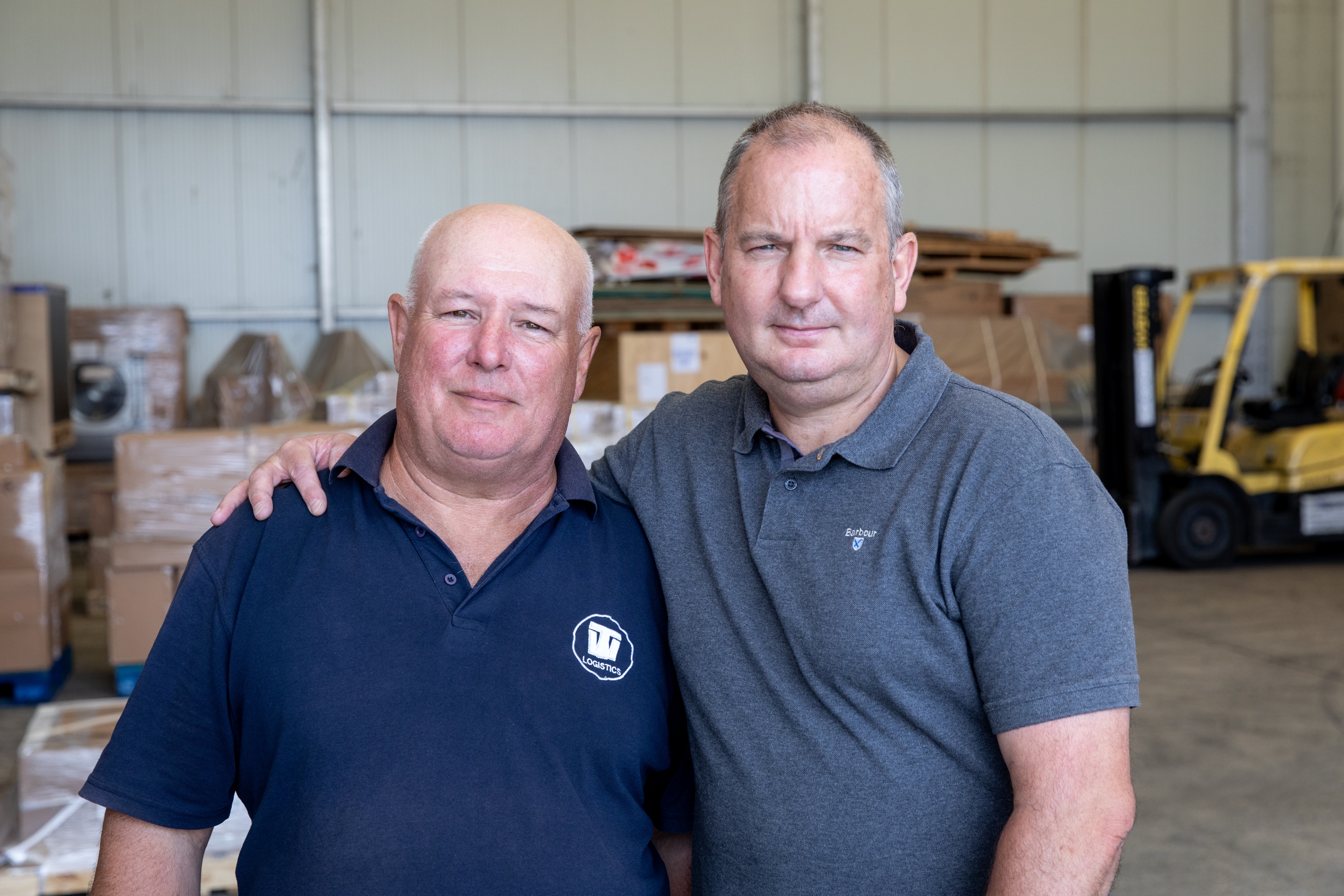 A photograph of Paul (left) and Mark (right) standing in a warehouse. Mark has his arm around Paul.