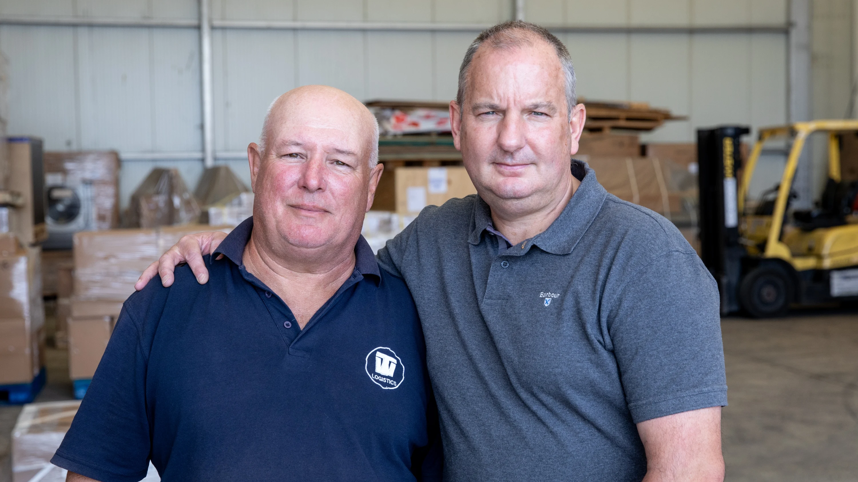 A photograph of Paul (left) and Mark (right) standing in a warehouse. Mark has his arm around Paul.
