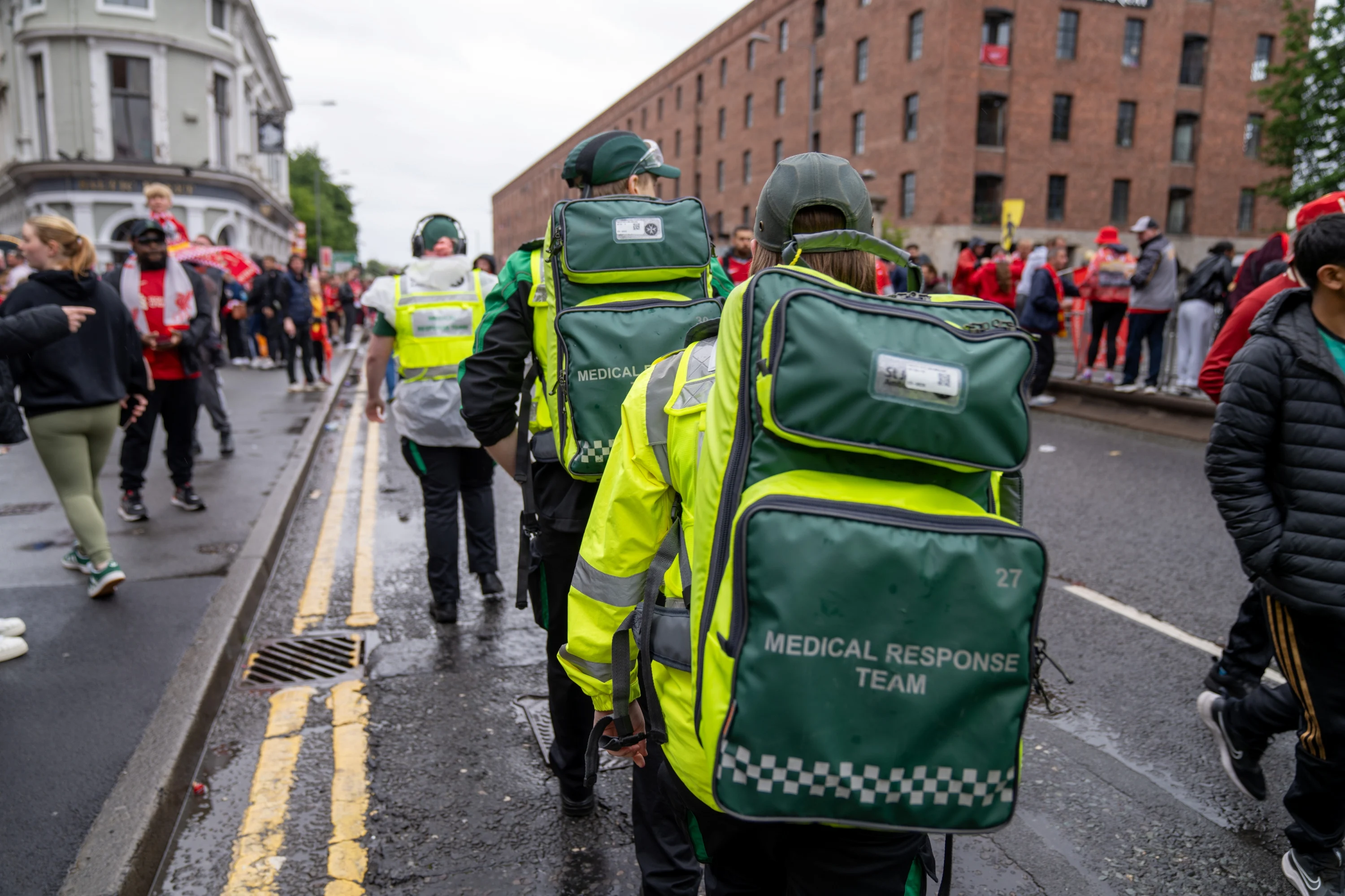 The back of three medical response team volunteers walking along a busy street. They are each wearing a large backpack.