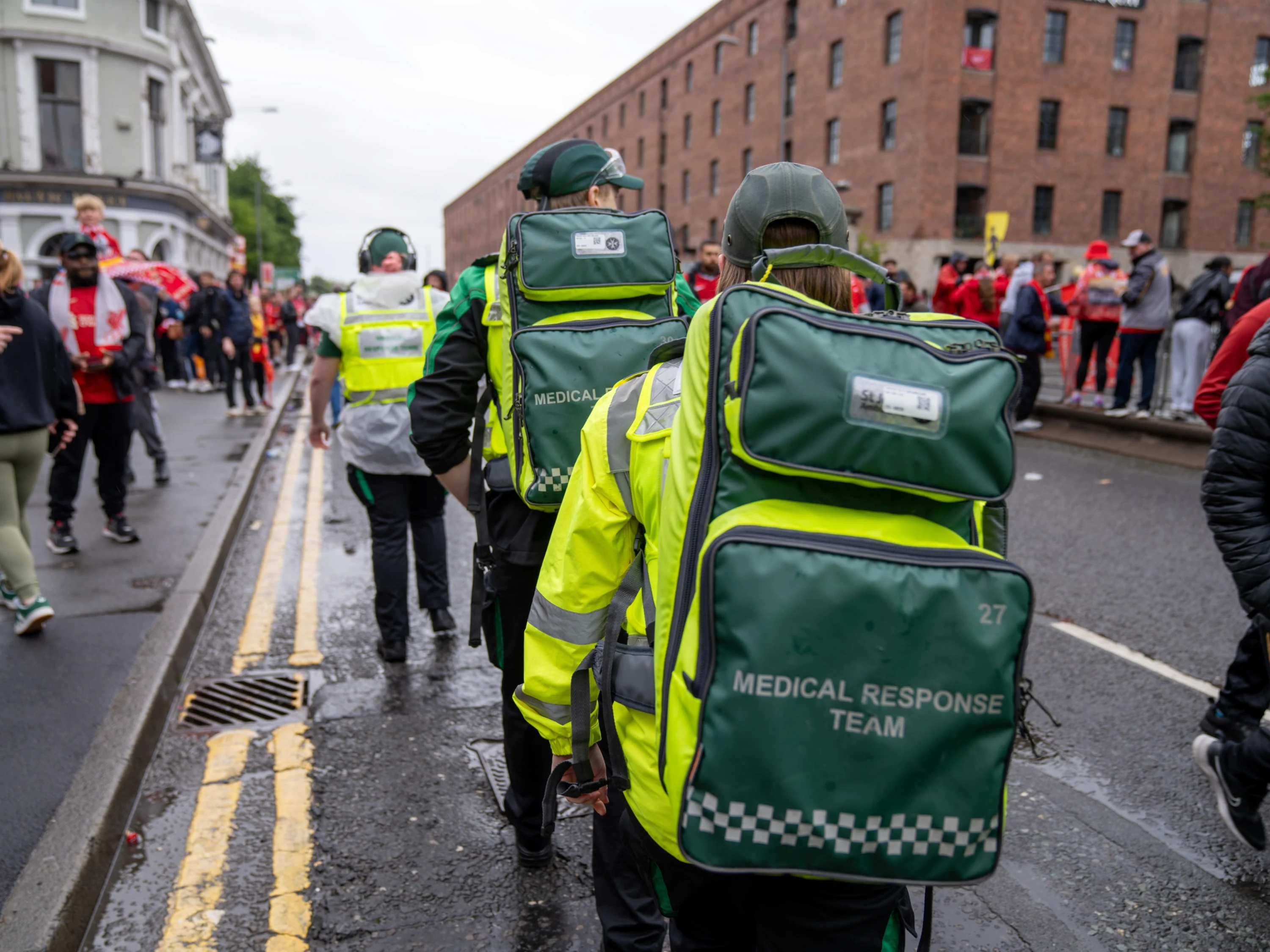 The back of three medical response team volunteers walking along a busy street. They are each wearing a large backpack.