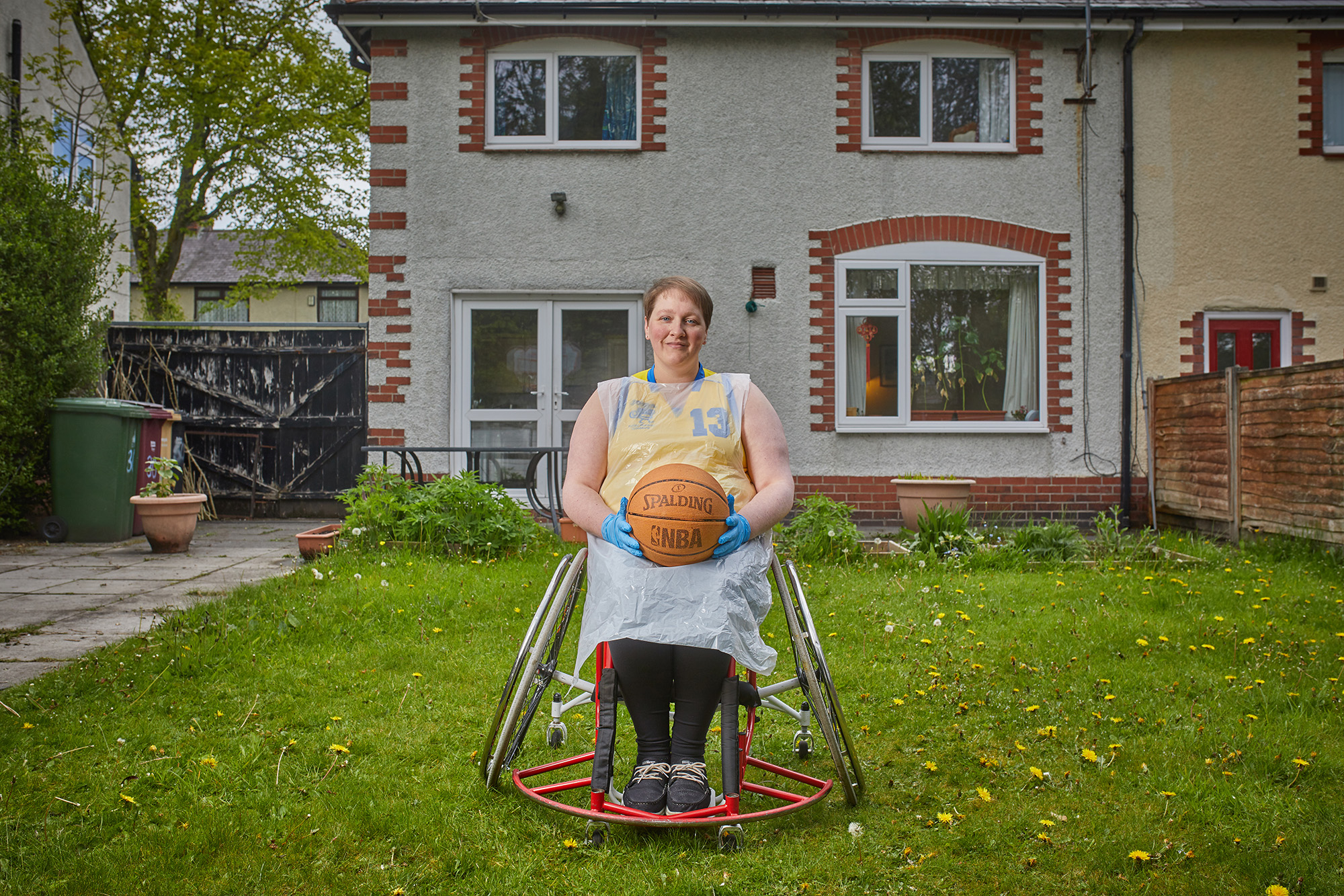 A photograph of Nia in her garden and wearing PPE. She is in a wheelchair and holding a basketball.