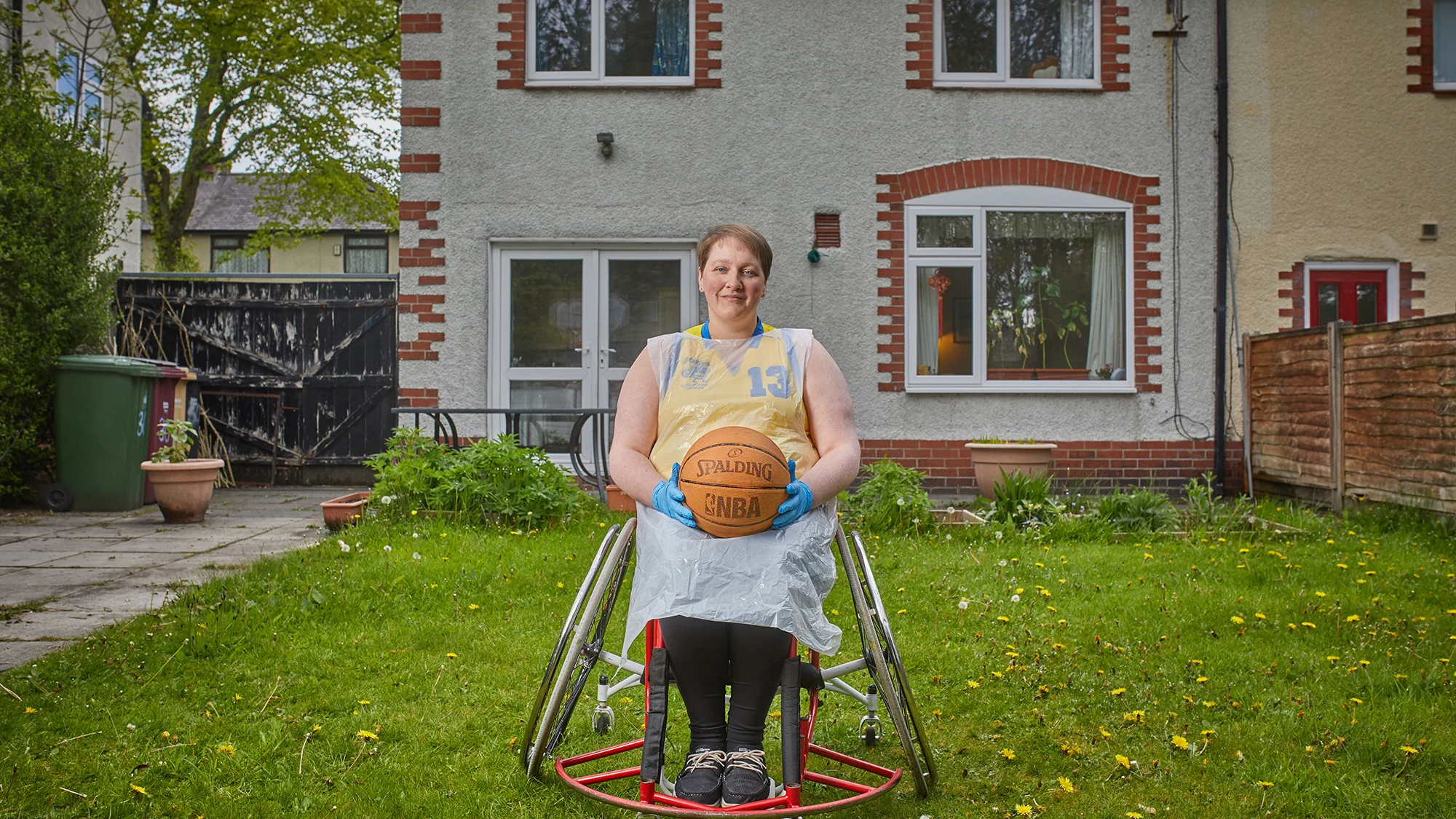 A photograph of Nia in her garden and wearing PPE. She is in a wheelchair and holding a basketball.