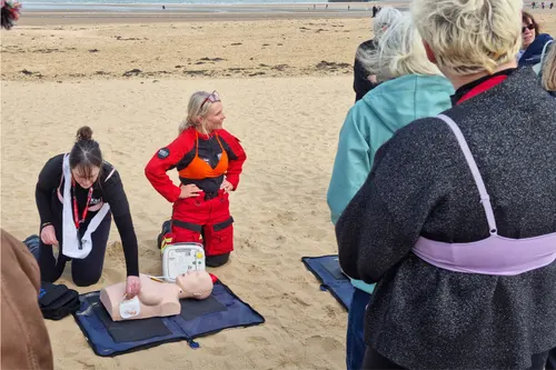 A photograph of women on a beach in Margate. The women are dressed and wearing their bras over their clothing. Two women in the centre of the image are performing a first aid demonstration on a female Manikin.