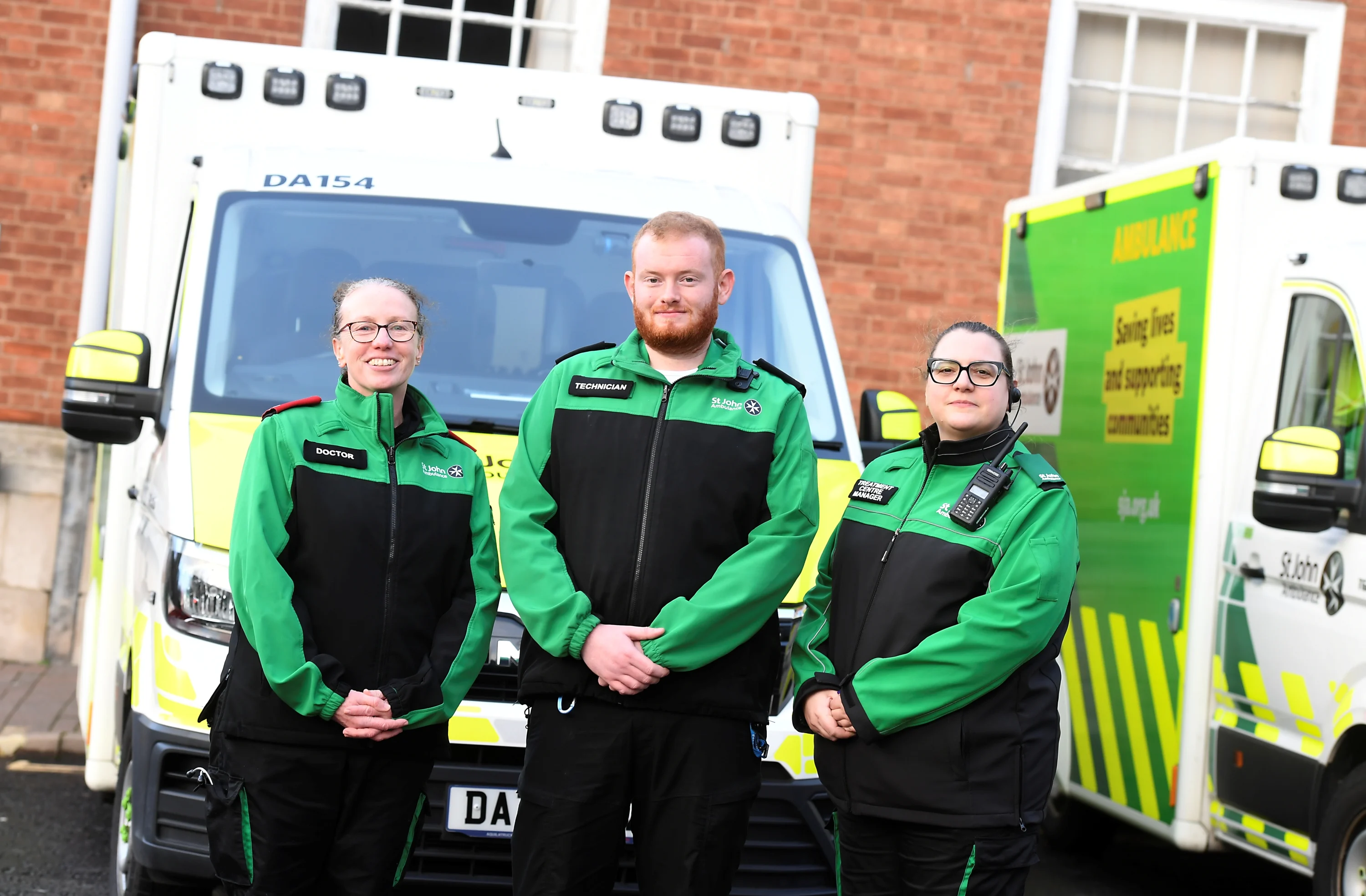 Three St John Ambulance volunteers stood in front of two treatment centre vehicles. One volunteer is wearing a 'Doctor' badge, another has a 'Technician' badge and another has a 'Treatment Centre Manager' badge. 