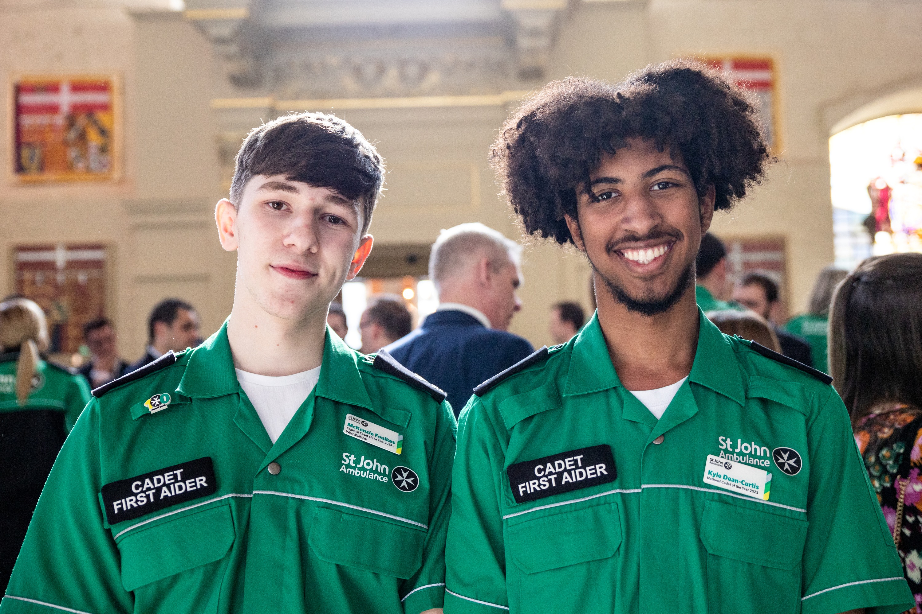 Two smiling Cadet First Aiders standing in a large old hall with lots of people standing around in the background.