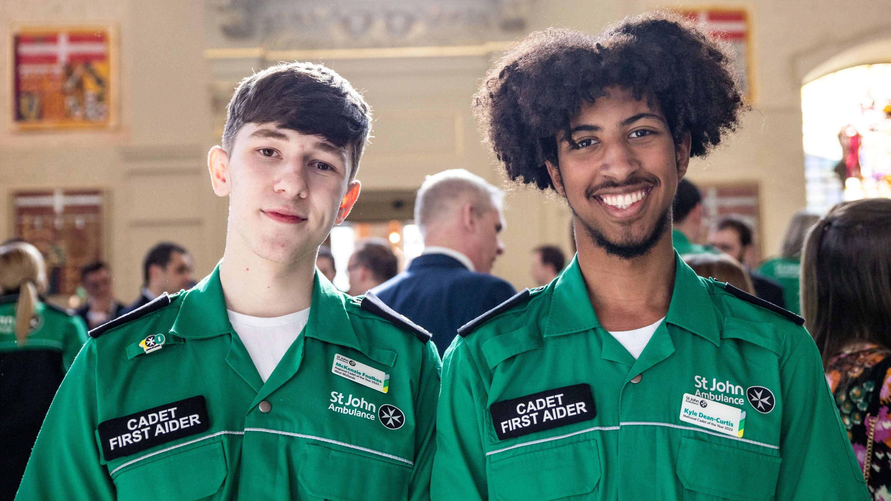 Two smiling Cadet First Aiders standing in a large old hall with lots of people standing around in the background.