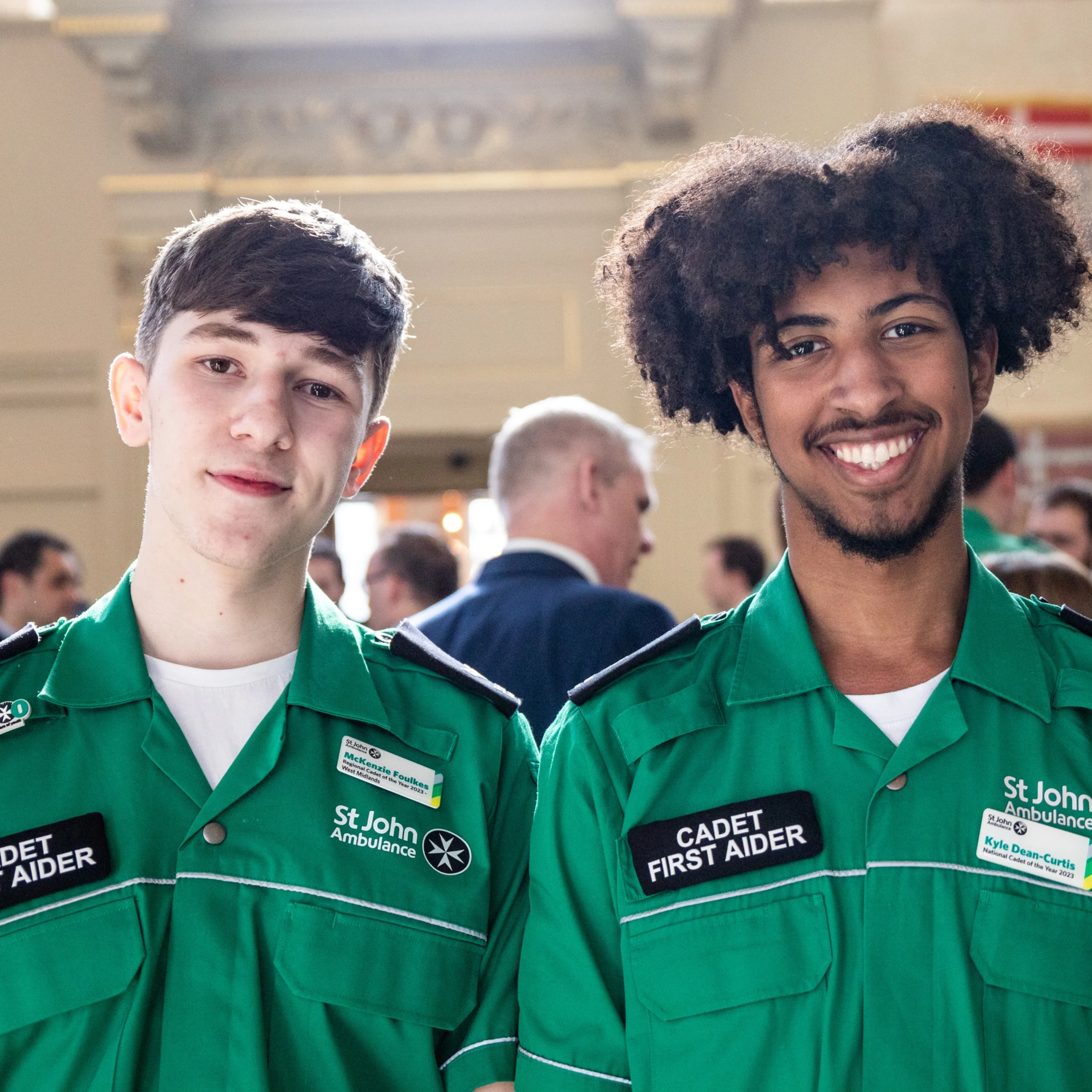 Two smiling Cadet First Aiders standing in a large old hall with lots of people standing around in the background.