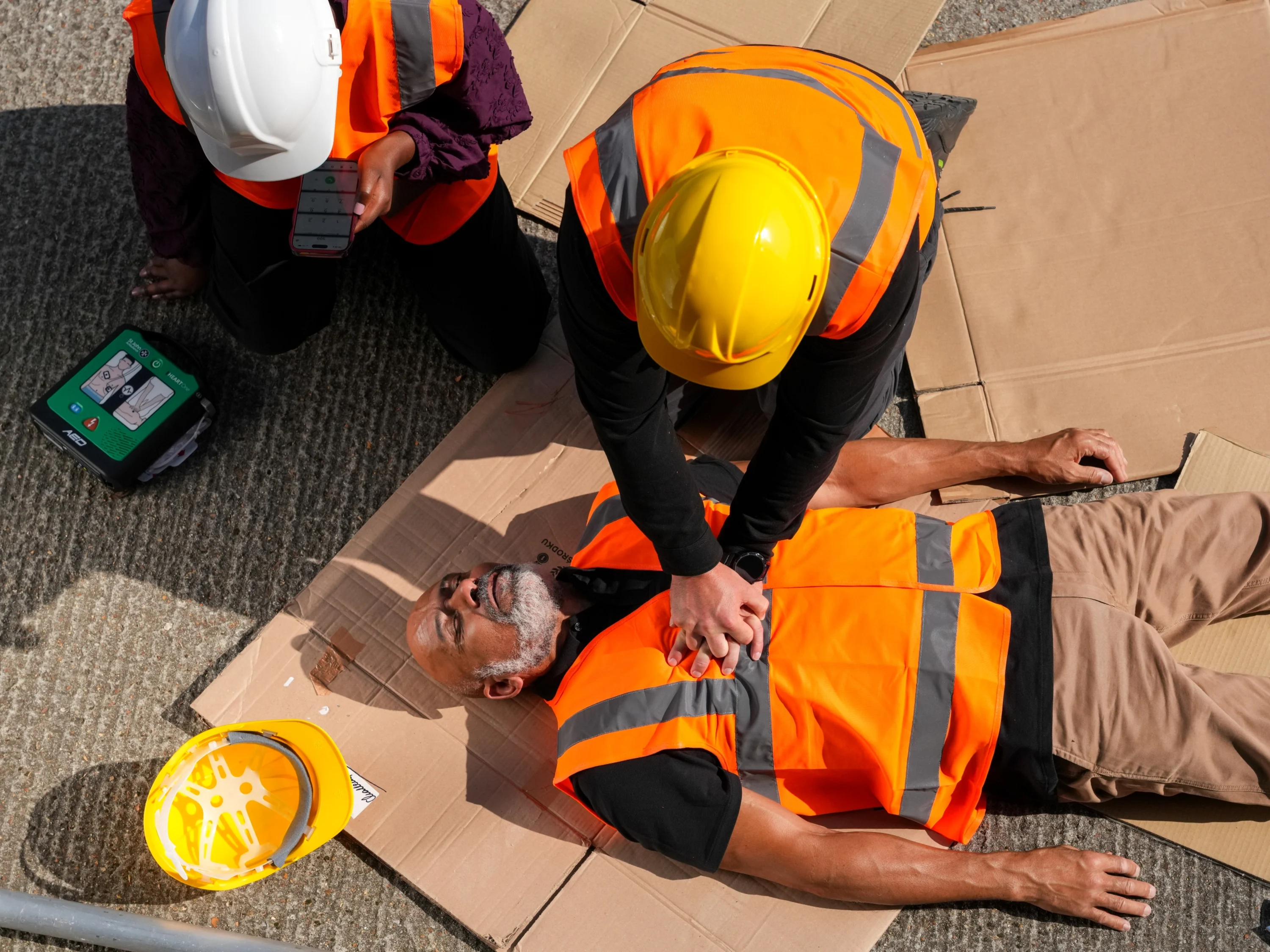An image of a first aider performing chest compressions on a man on a building site. There is a defibrillator alongside them.