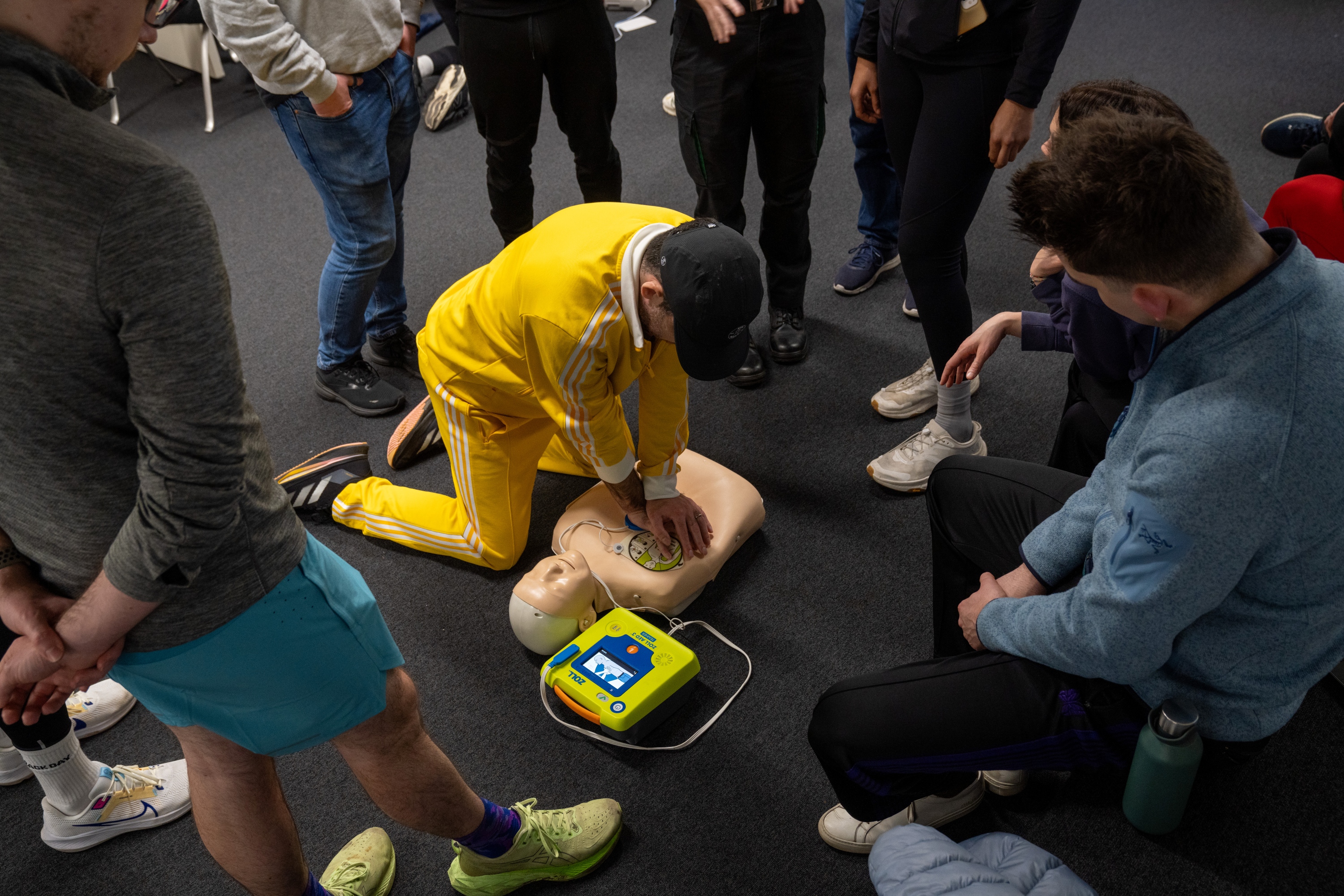 A photograph of a man in a bright yellow tracksuit and black baseball cap practising chest compressions on a Manikin. A group of people in running gear gather around and watch him.