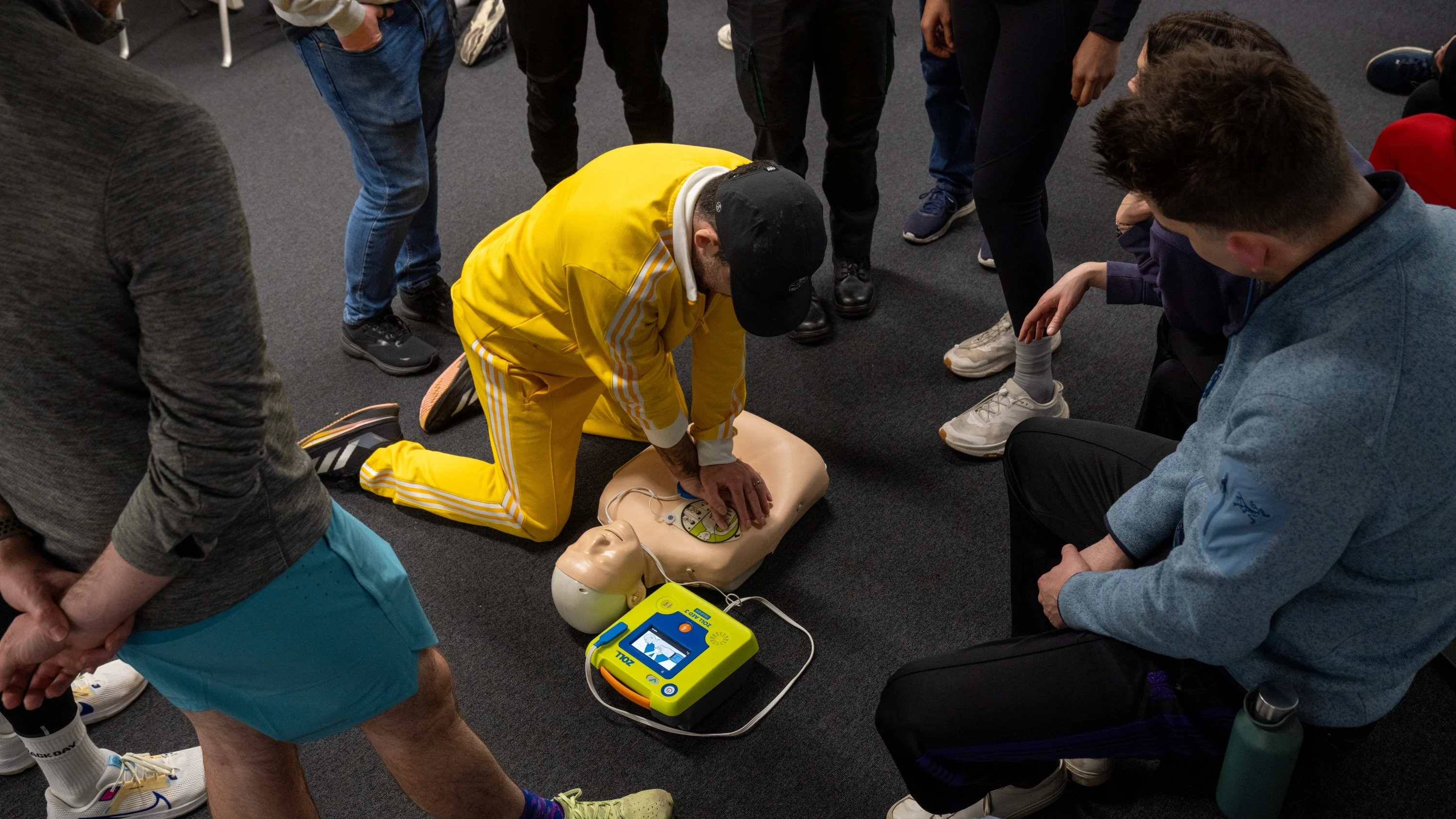 A photograph of a man in a bright yellow tracksuit and black baseball cap practising chest compressions on a Manikin. A group of people in running gear gather around and watch him.