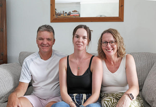 A photograph of Gary, Olivia and Karen Pitfield (left to right), sitting on the sofa in their living room and smiling at the camera.
