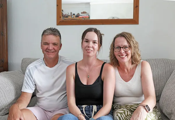 A photograph of Gary, Olivia and Karen Pitfield (left to right), sitting on the sofa in their living room and smiling at the camera.