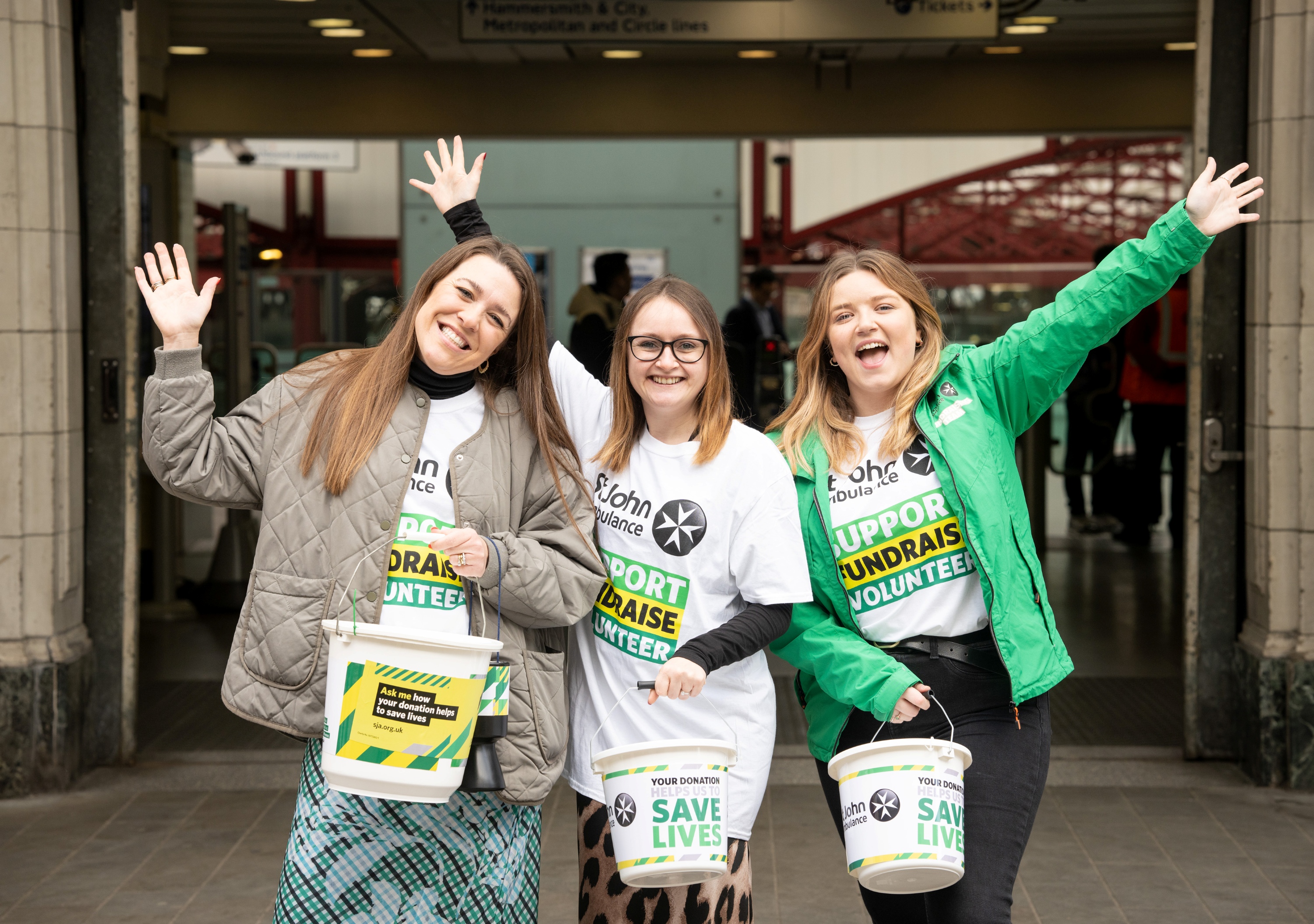 Three people wearing St John Ambulance fundraising t-shirts, smiling, with their arms in the air, in front of large glass doors. They're also each holding a large collection bucket. 