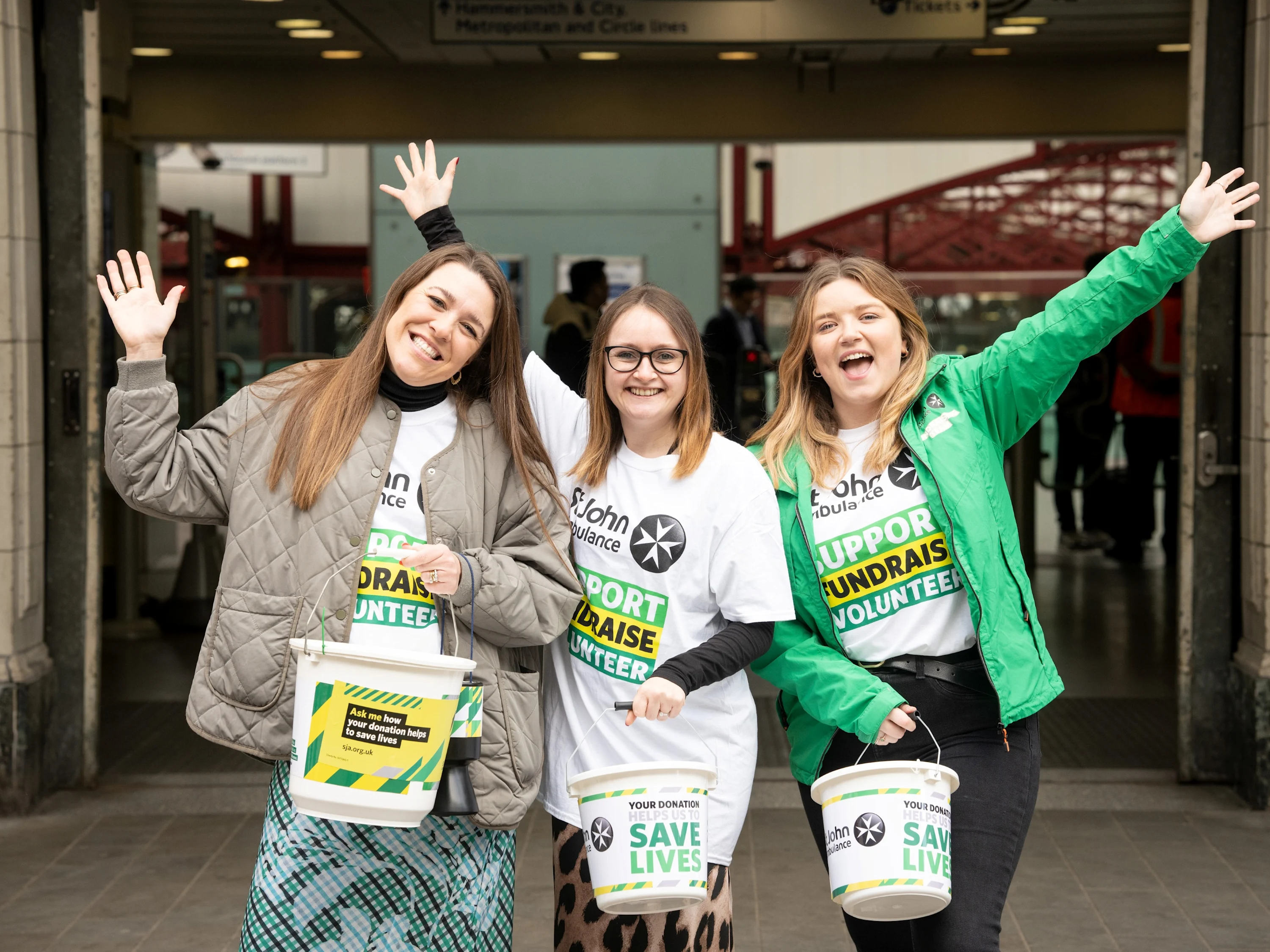 Three people wearing St John Ambulance fundraising t-shirts, smiling, with their arms in the air, in front of large glass doors. They're also each holding a large collection bucket.