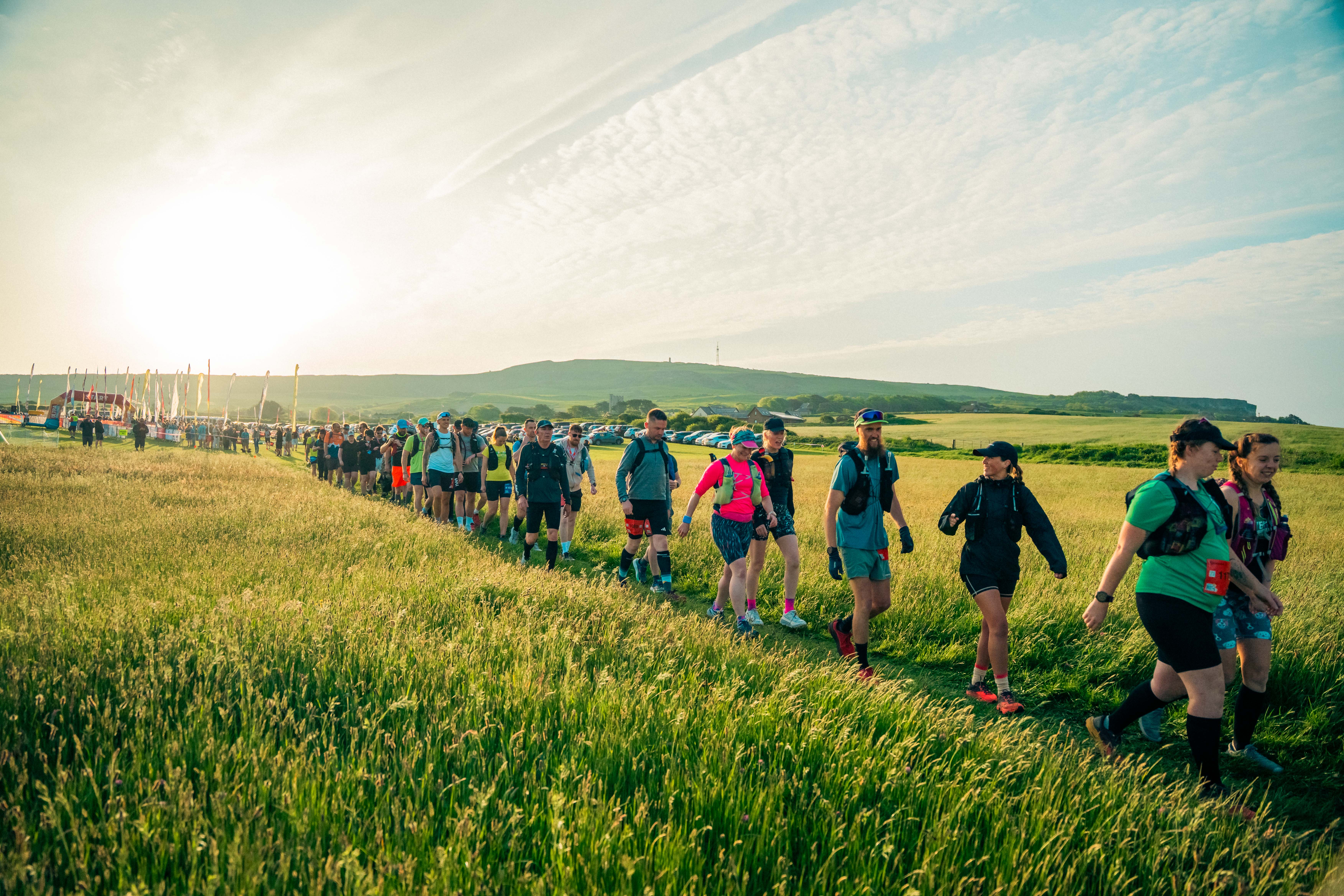 A large group of people wearing active wear walking across a field in a line with the sun setting behind them. In the distance are large flags on poles and an arch. 