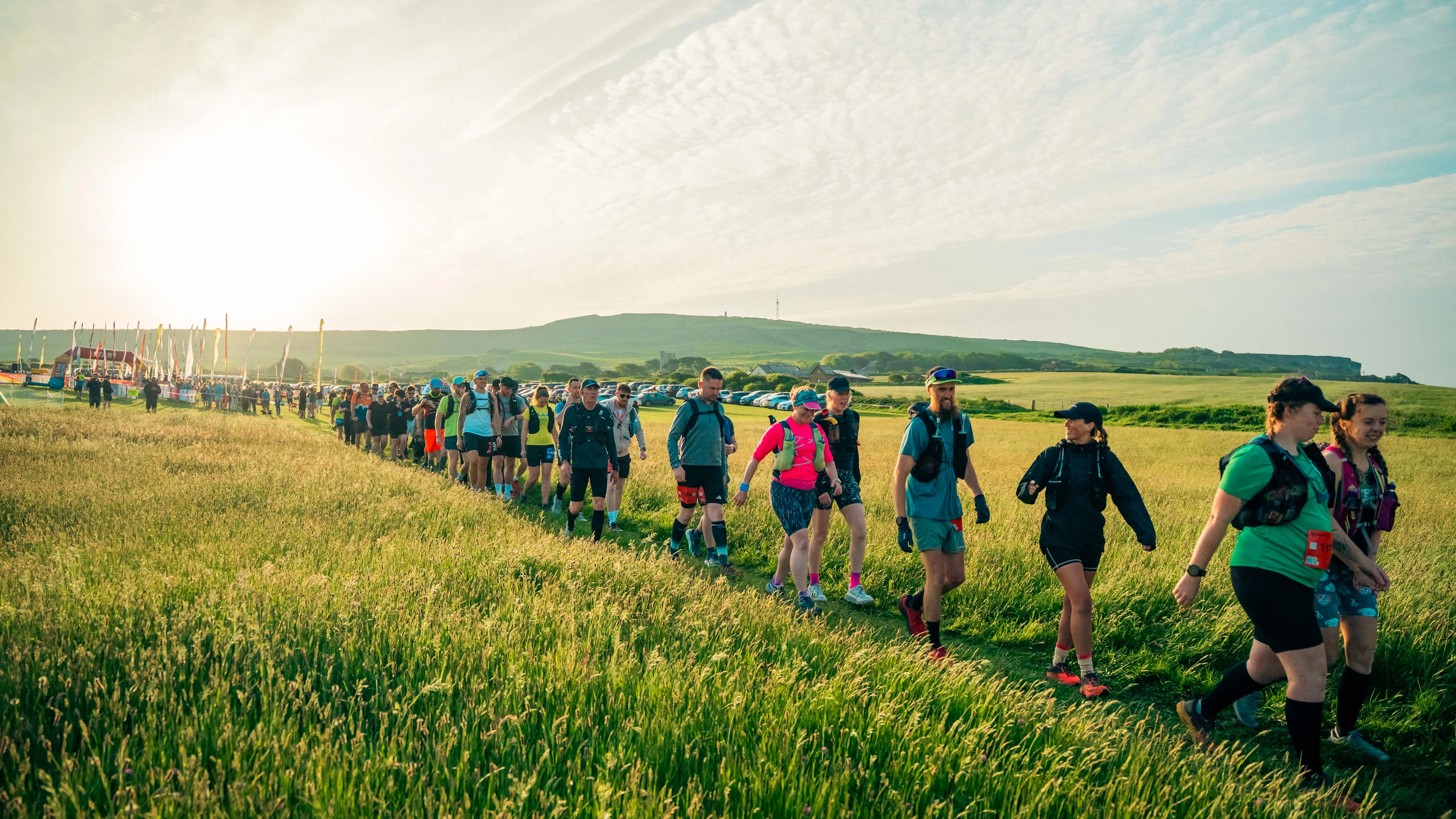 A large group of people wearing active wear walking across a field in a line with the sun setting behind them. In the distance are large flags on poles and an arch.