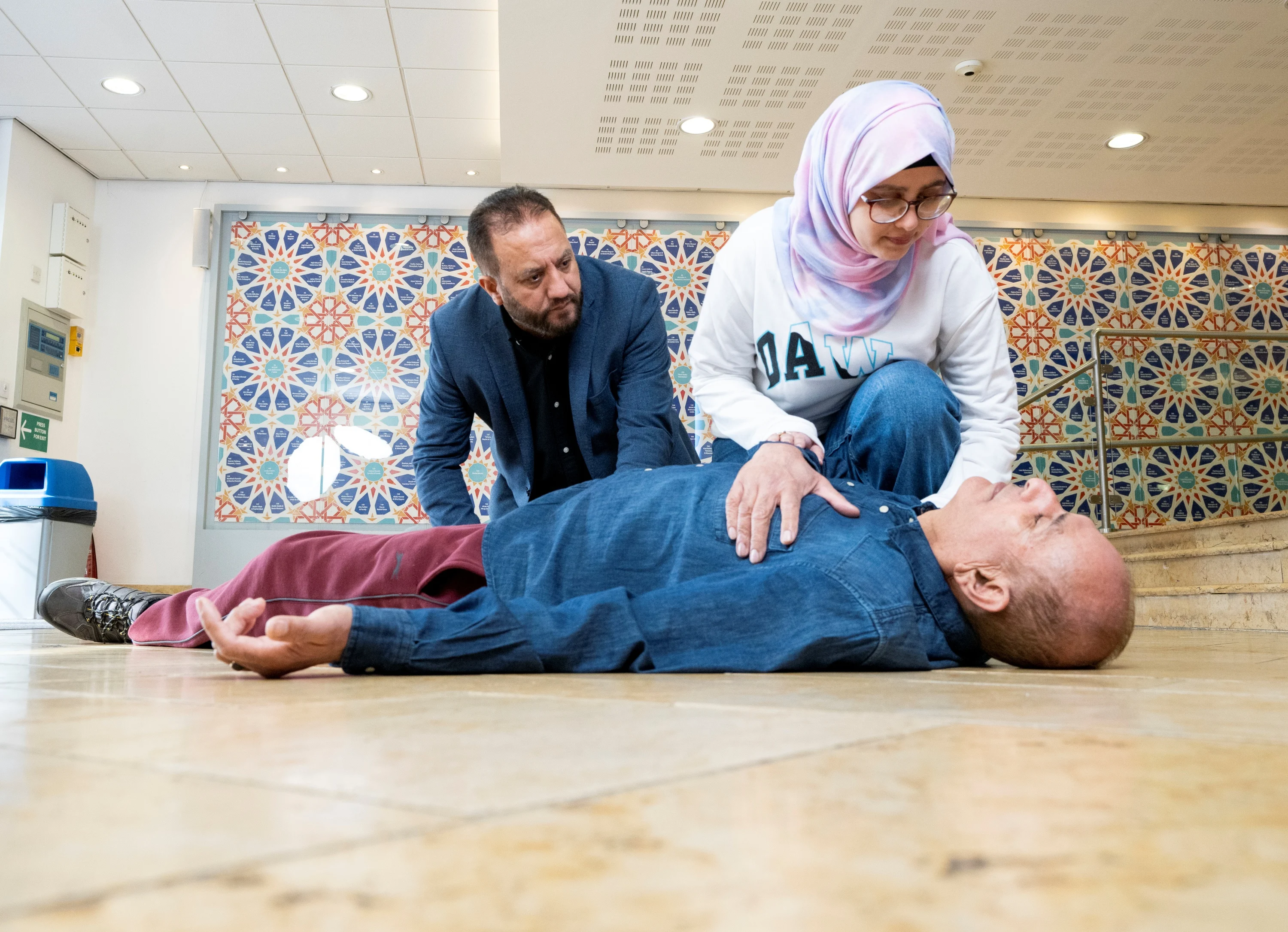 Two people kneeling down on the floor. One is leaning over with their hand on the chest of someone lying down on the floor with their eyes closed. 