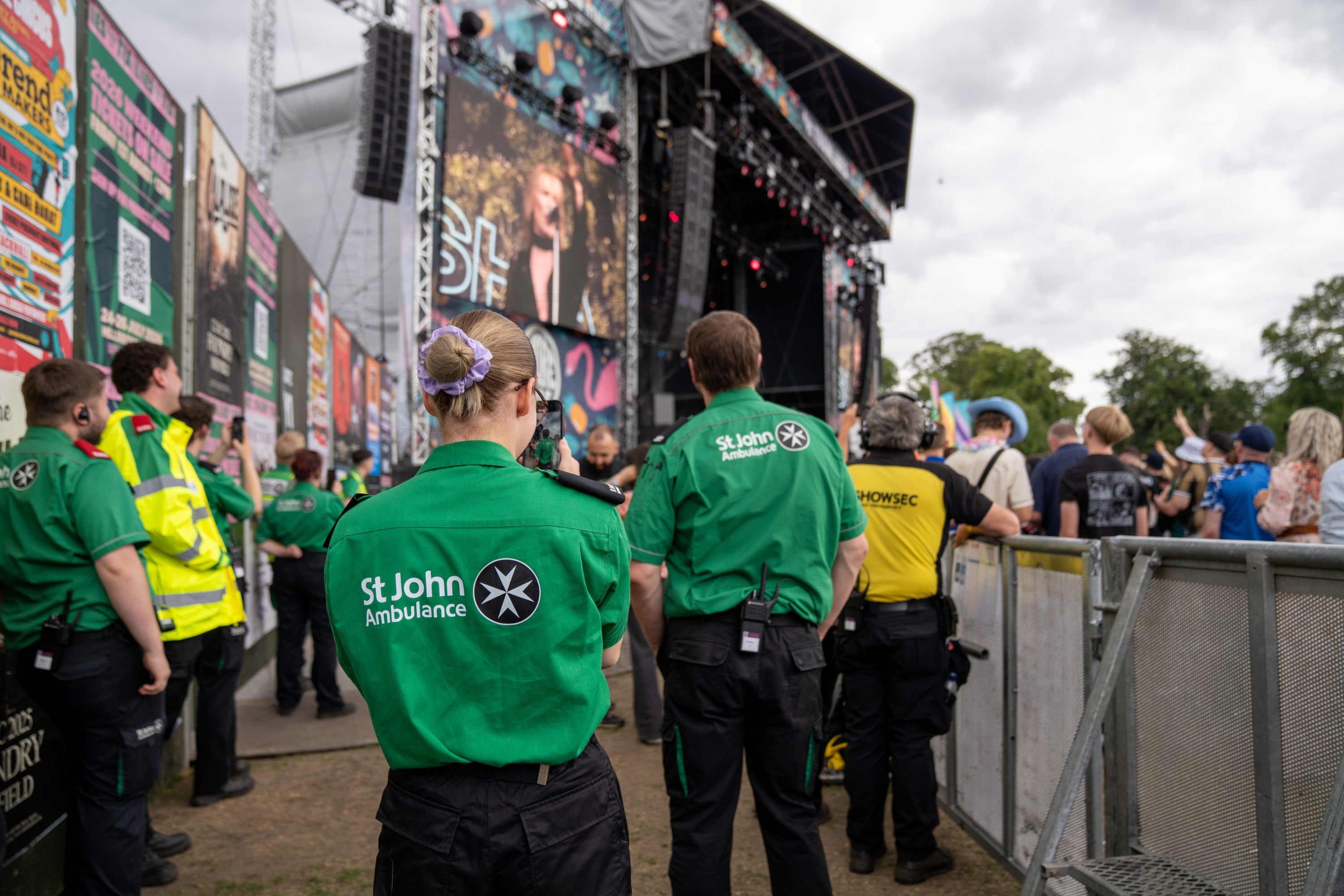 The back of two volunteers looking at a large concert stage outdoors. There are other first aid volunteers standing around and the audience also looking at the stage behind a barrier. 