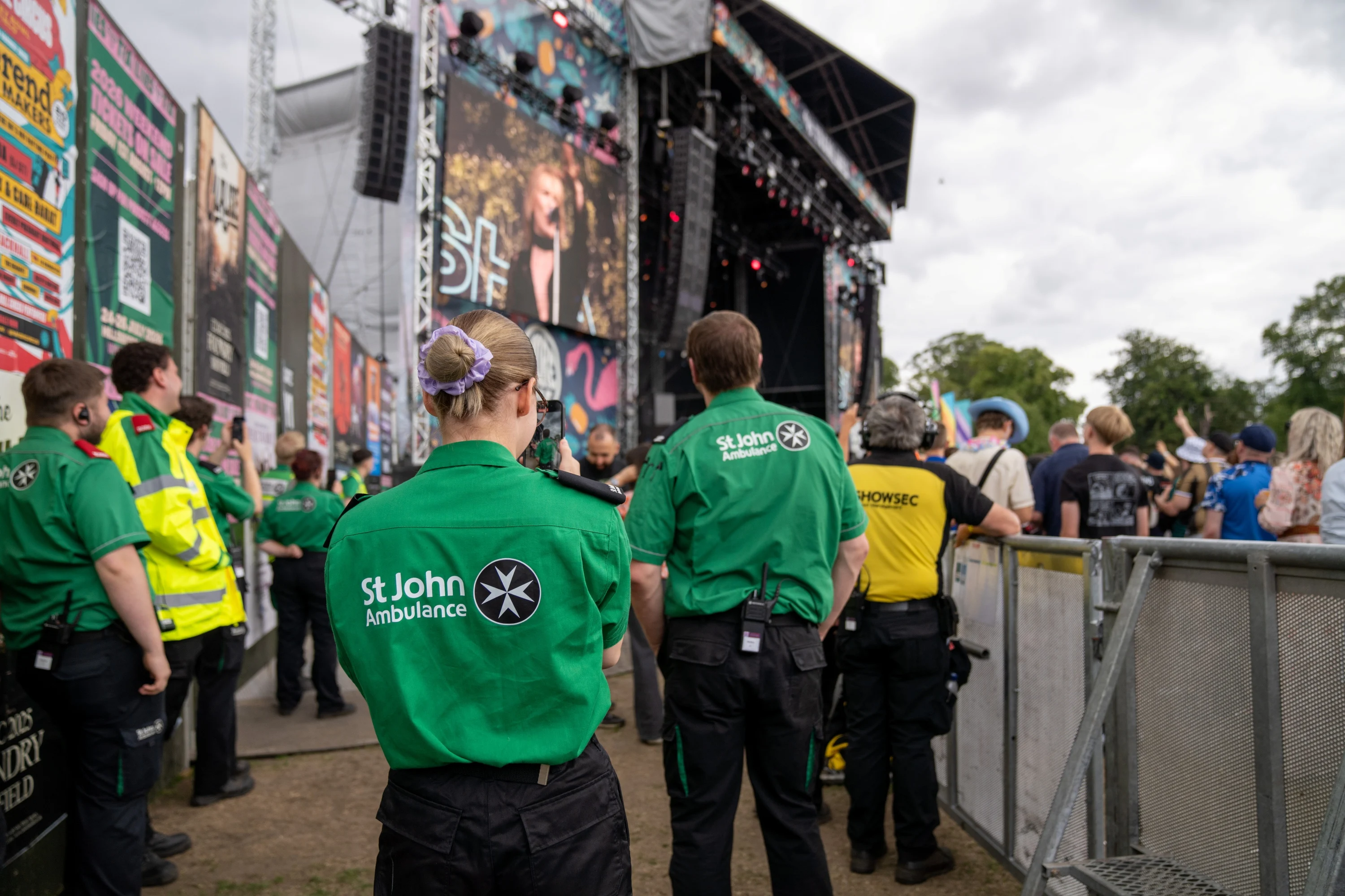 The back of two volunteers looking at a large concert stage outdoors. There are other first aid volunteers standing around and the audience also looking at the stage behind a barrier.