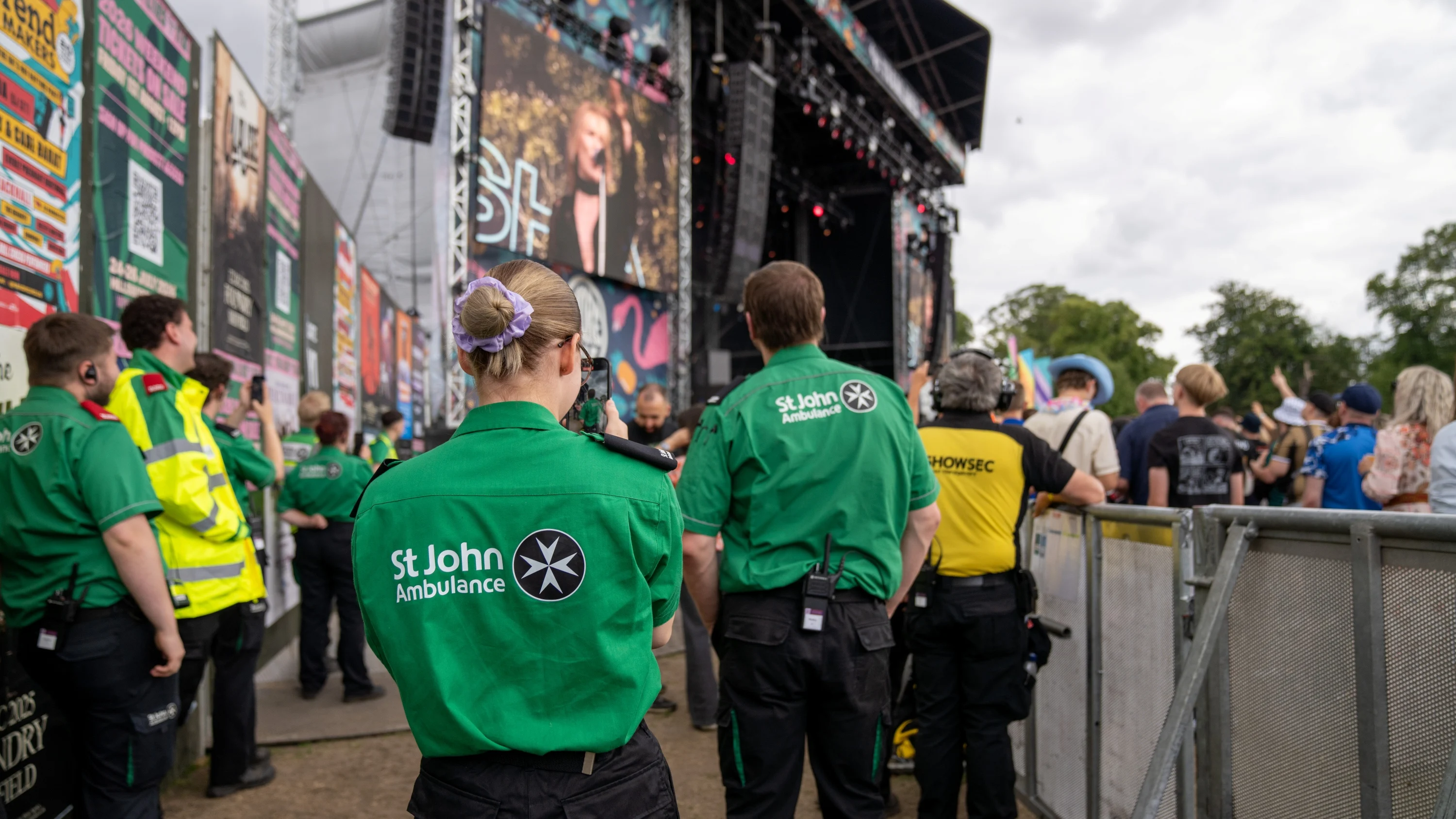 The back of two volunteers looking at a large concert stage outdoors. There are other first aid volunteers standing around and the audience also looking at the stage behind a barrier.