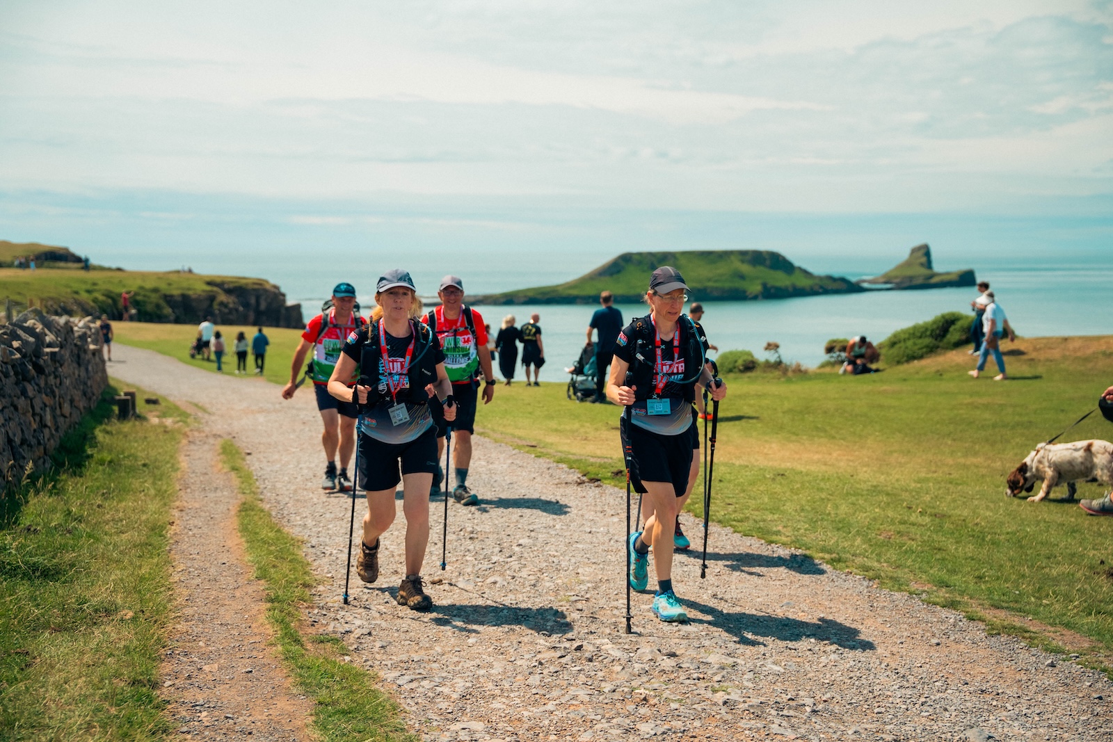A group of walkers with walking sticks and backpacks walking along a coastal footpath on the Gower Peninsula. It's a sunny day and the sea is in the background.