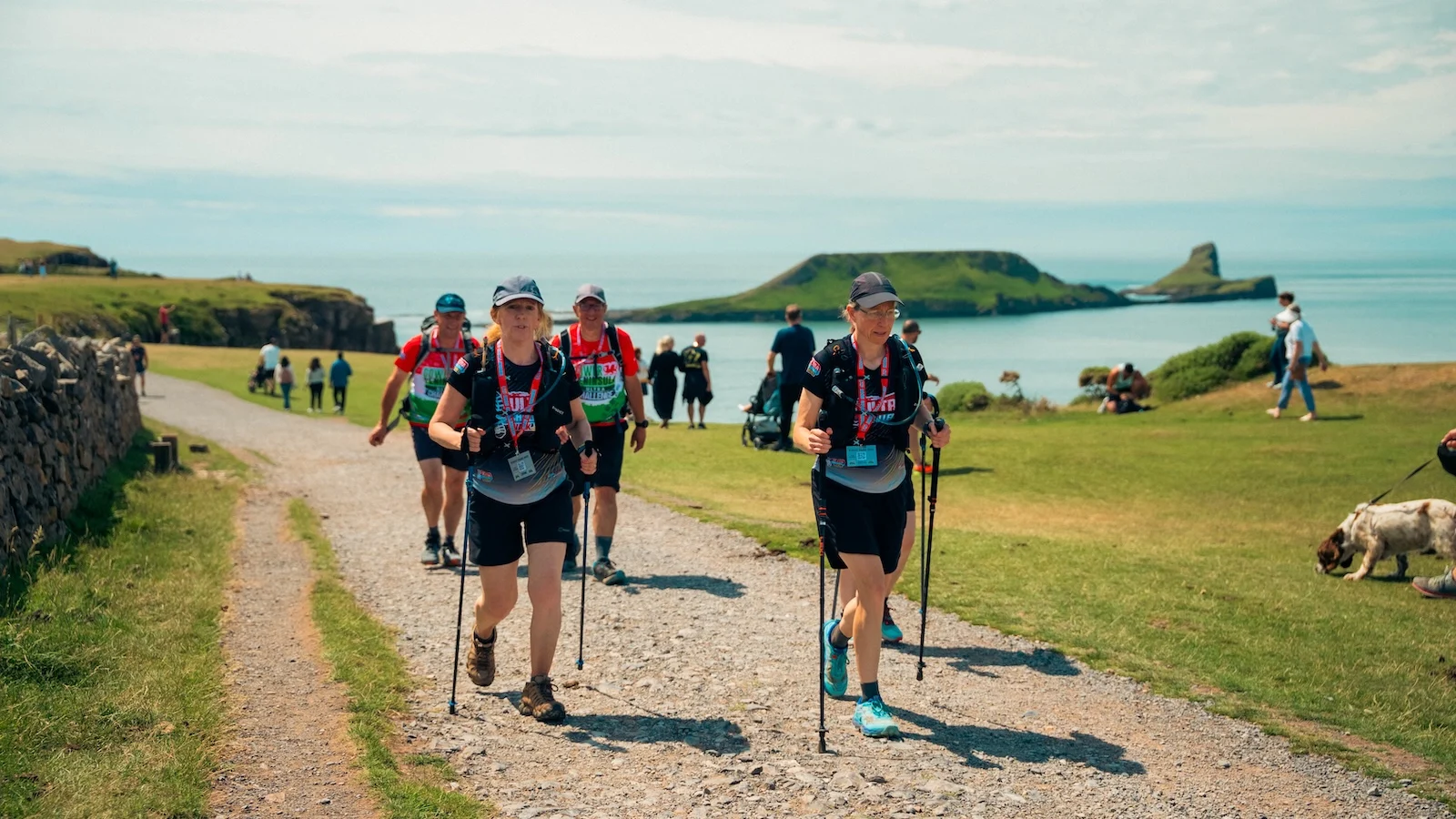 A group of walkers with walking sticks and backpacks walking along a coastal footpath on the Gower Peninsula. It's a sunny day and the sea is in the background.
