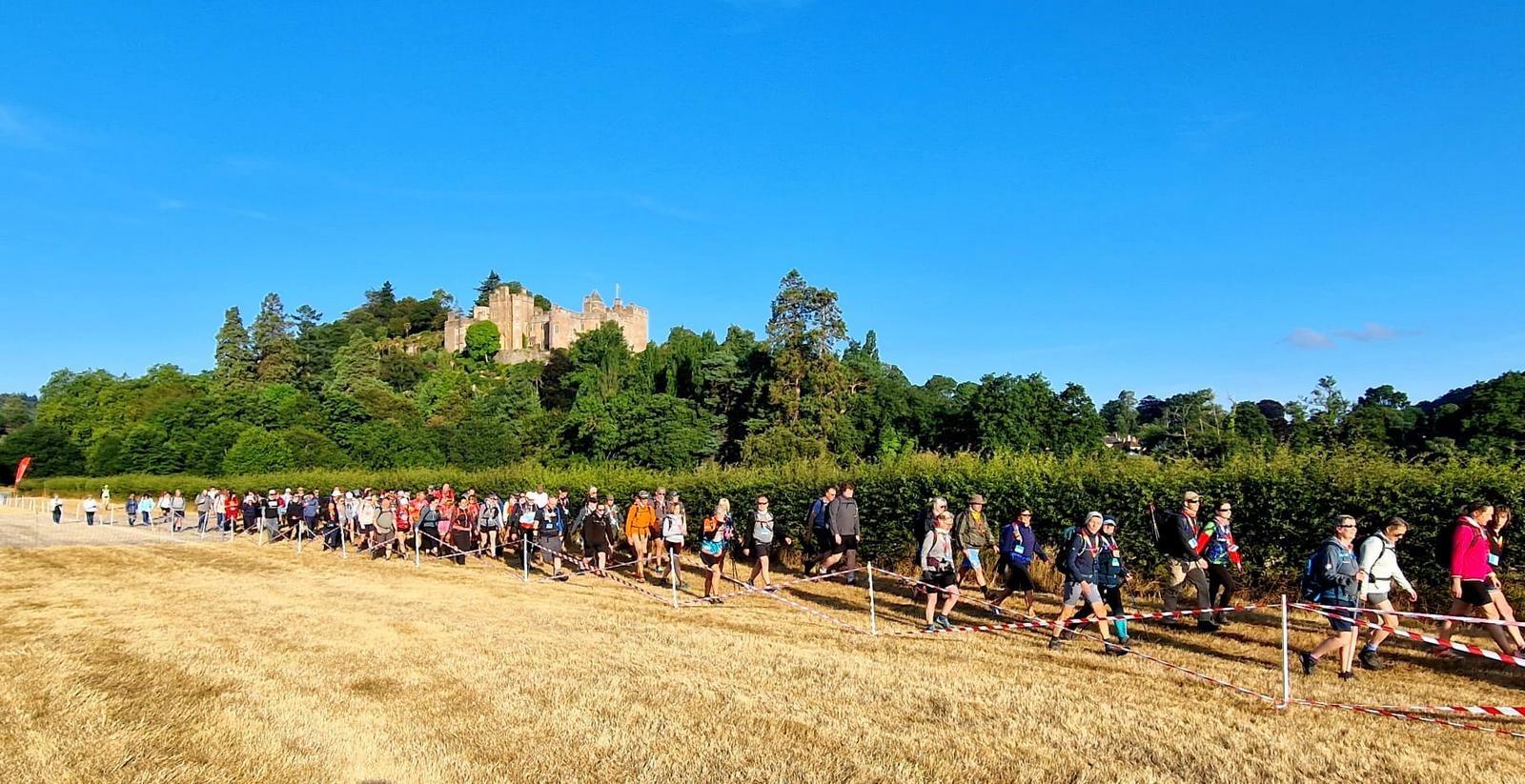 A large group of people walking along the edge of a field with a path cordoned off. There is a large castle and woodland in the background.