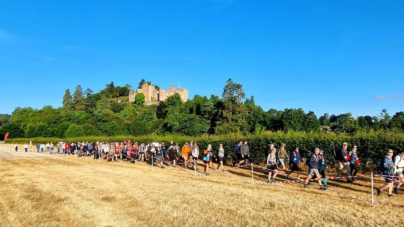 A large group of people walking along the edge of a field with a path cordoned off. There is a large castle and woodland in the background.