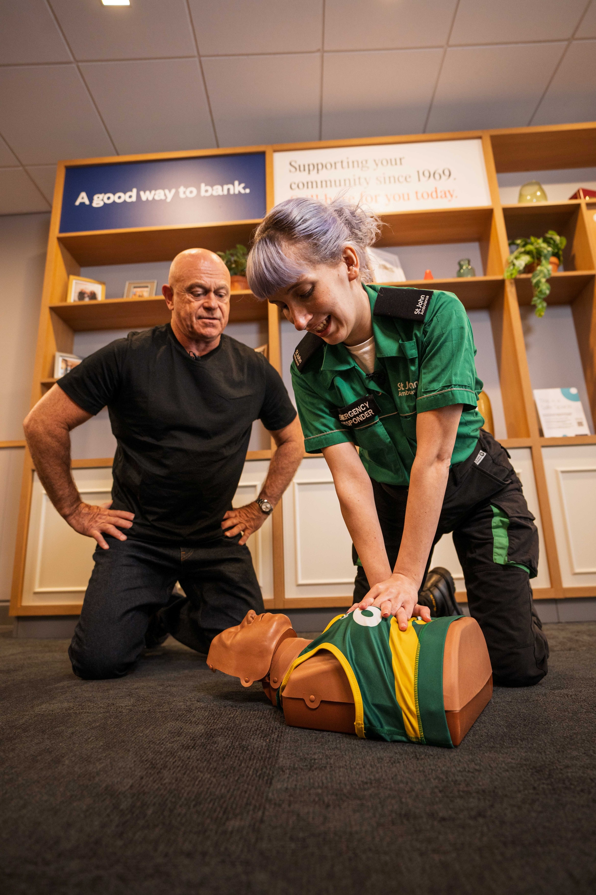 Ross Kemp with St John Ambulance first aid trainer Flora, kneeling on the floor around a CPR manikin. Flora has her hands on the manikin doing chest compressions. Ross Kemp is observing.