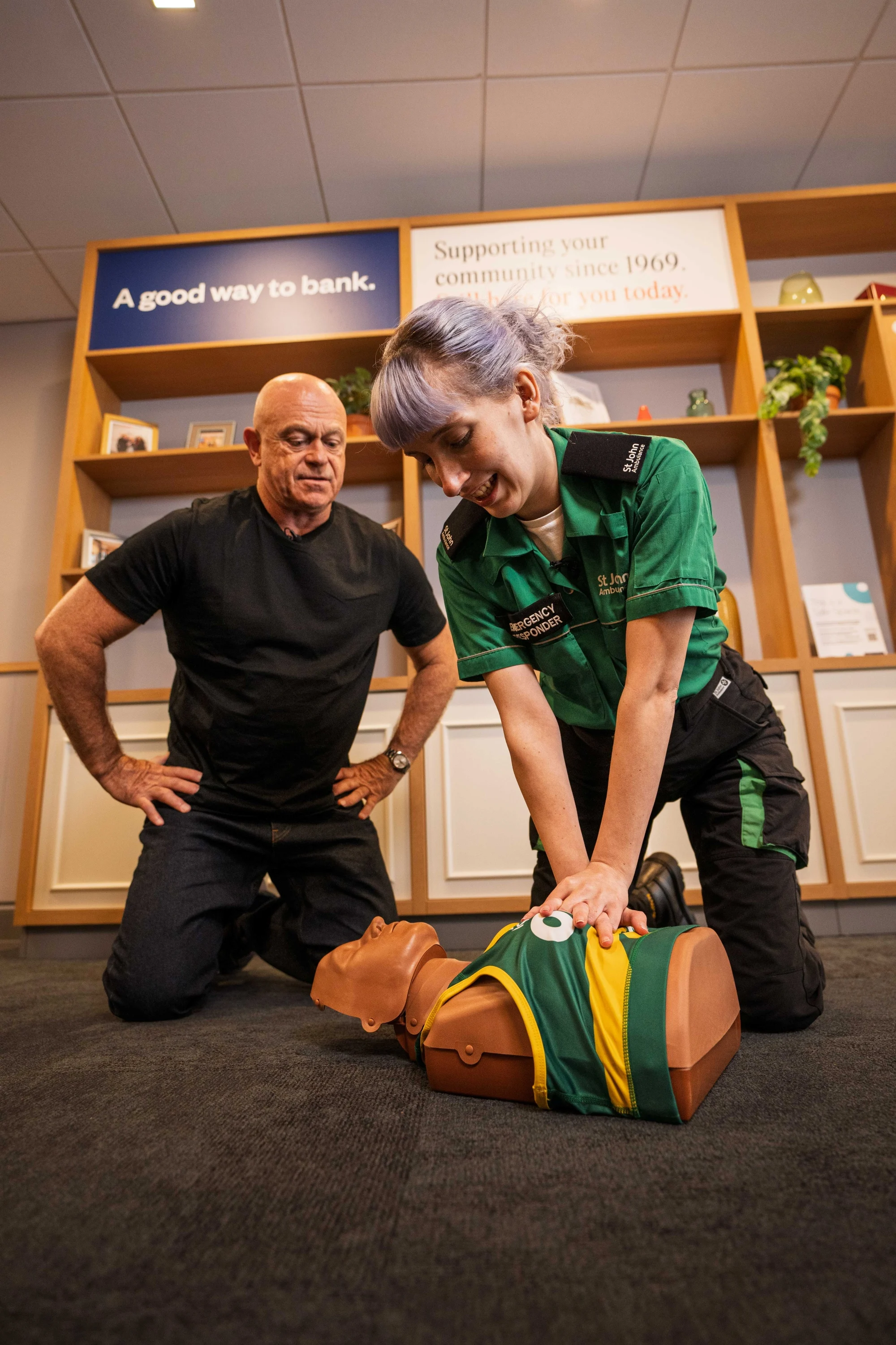 Ross Kemp with St John Ambulance first aid trainer Flora, kneeling on the floor around a CPR manikin. Flora has her hands on the manikin doing chest compressions. Ross Kemp is observing.