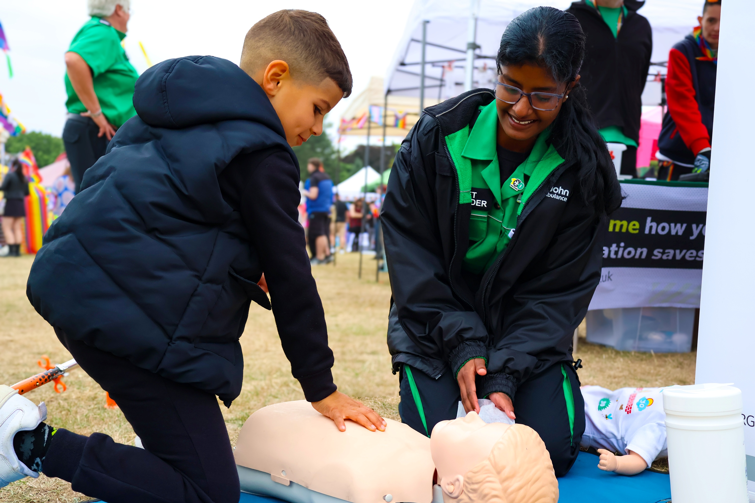 A Cadet First Aider and child kneeling on the floor at a busy outdoor event. The child is doing a chest compression on a CPR mannequin. 