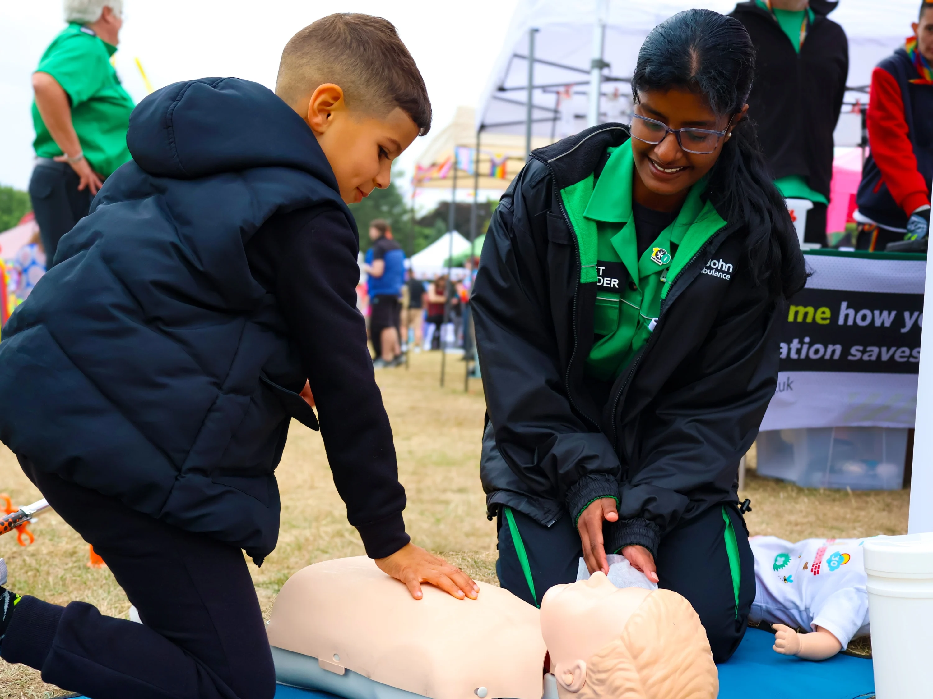 A Cadet First Aider and child kneeling on the floor at a busy outdoor event. The child is doing a chest compression on a CPR mannequin.